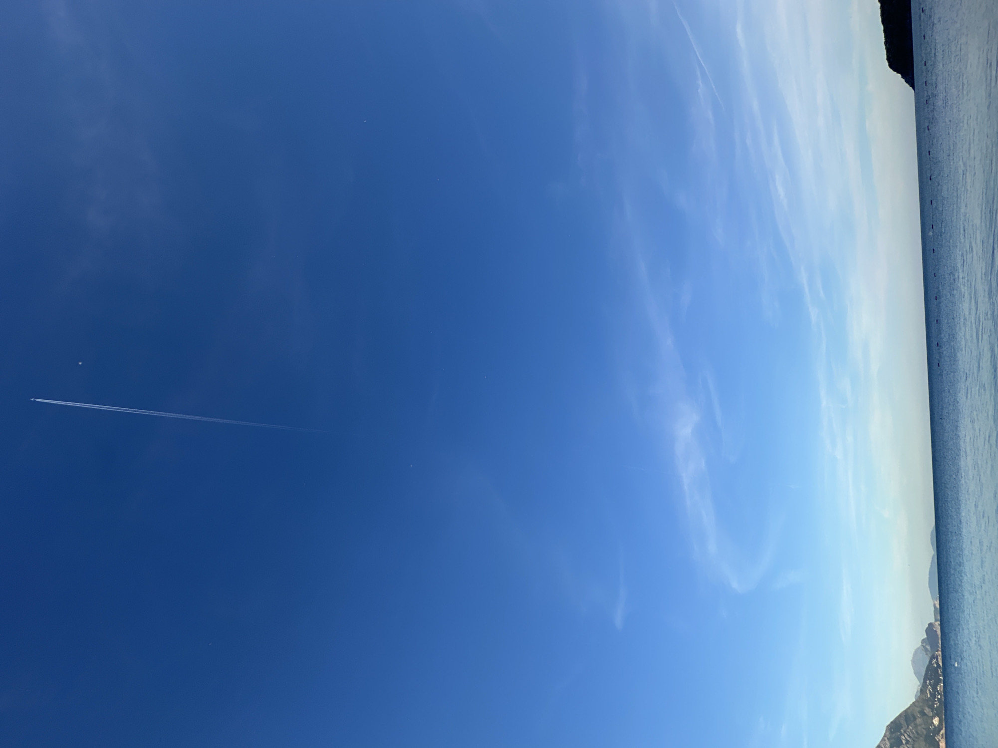 The image depicts a daytime scene in or near the city of Prijevor, Montenegro. The sky is predominantly blue, with some cirrus clouds visible towards the right side of the frame. A contrail from a jet aircraft stretches across the sky from left to right. The rightmost edge of the frame shows the edge of what appears to be water and land/mountains. No people are visible.