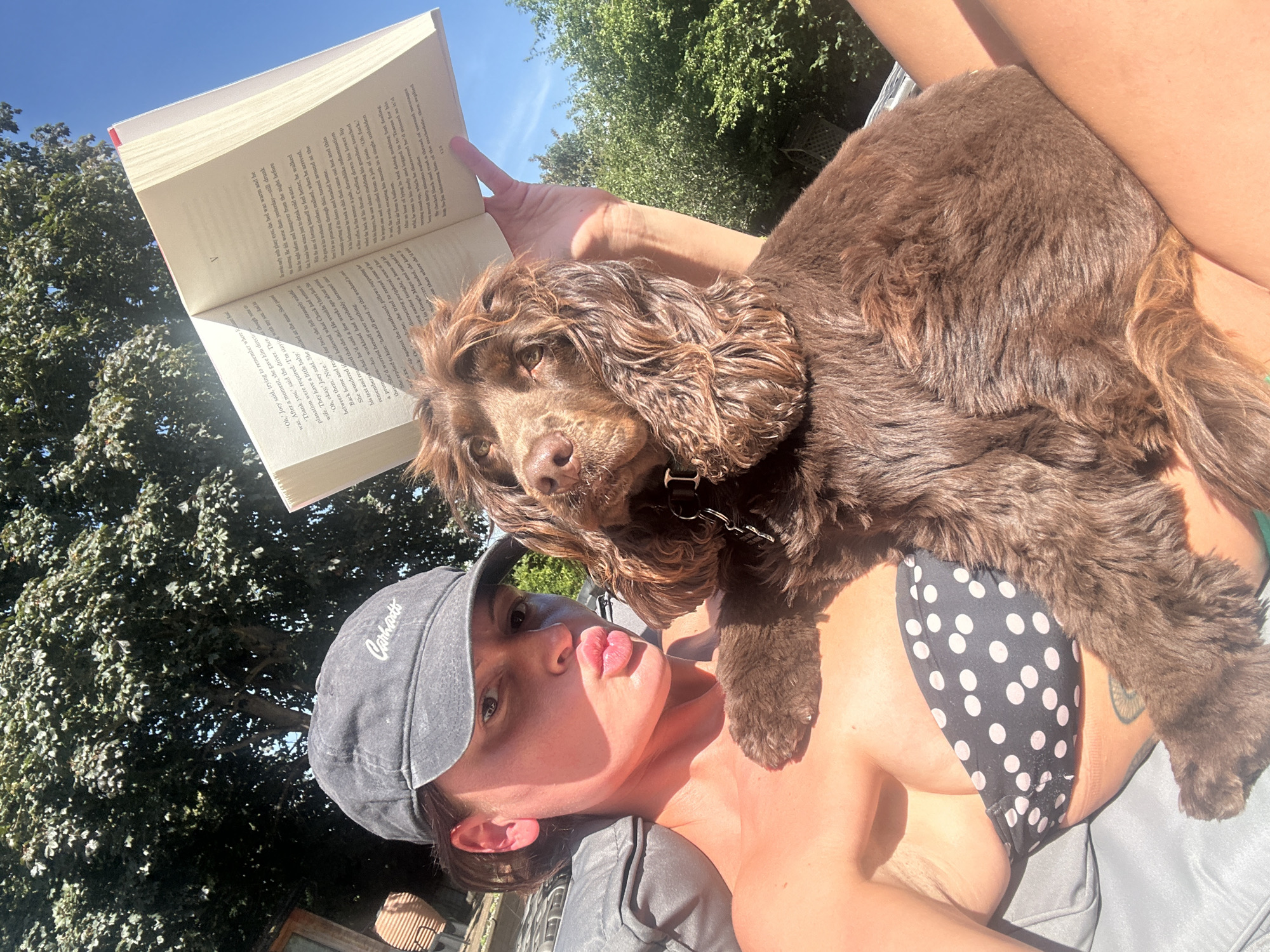 A person wearing a black baseball cap and a black and white polka dot bikini top is sitting outdoors, holding a book open with their left hand and a brown dog on their lap with their right arm. The person has their lips pursed in a "duck face" pose. The dog is looking towards the camera. The background shows trees and a bright blue sky. The scene appears to be a sunny day in a garden. There is no information in the image to suggest it is located in Tunbridge Wells, United Kingdom.