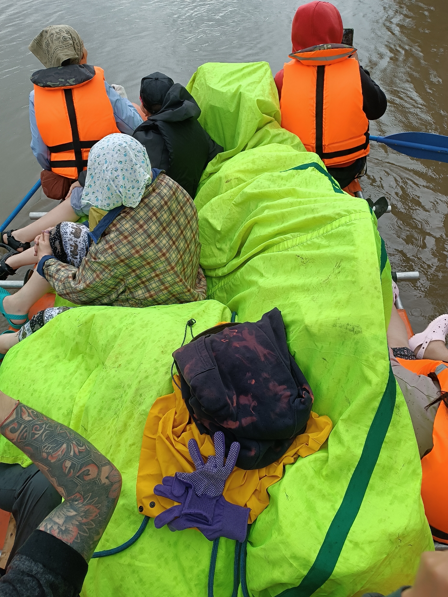 The image shows a group of people on a raft in murky water, likely on the Arkhara River near Idrisovo, Russia. The people are wearing life vests and head coverings. The raft is covered with bright green tarps, and there are various items of clothing and a pair of purple gloves on top of the tarps. The water is choppy, indicating movement, and a blue paddle is visible in the upper right corner of the image, suggesting the raft is being paddled. The overall scene conveys an outdoor activity, possibly a rafting trip.