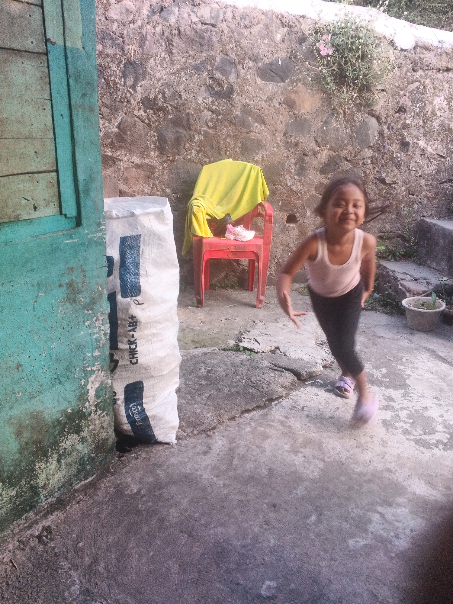 A young girl is in motion, captured mid-stride on a concrete ground. She is wearing a light-colored tank top and dark pants, with light-colored sandals on her feet. Her face is visible, and she appears to be smiling, with her hair slightly blurred from her movement. The scene takes place outdoors, with a rough stone wall in the background and a wooden structure on the left side of the frame, painted teal and showing signs of wear. To the right of the girl, there is a bright red plastic chair covered with a yellow cloth, and a pair of white and pink shoes placed on the seat. A large, white sack with blue lettering is visible on the left, partially obscuring the teal structure. A potted plant is also visible in the background. The lighting suggests it is daytime, with sunlight illuminating the scene. The overall setting appears to be a simple, possibly residential outdoor area in Keji, Indonesia. Visible text on the sack includes "CHICK-AB+ P" and what appears to be a logo or partial text at the bottom.