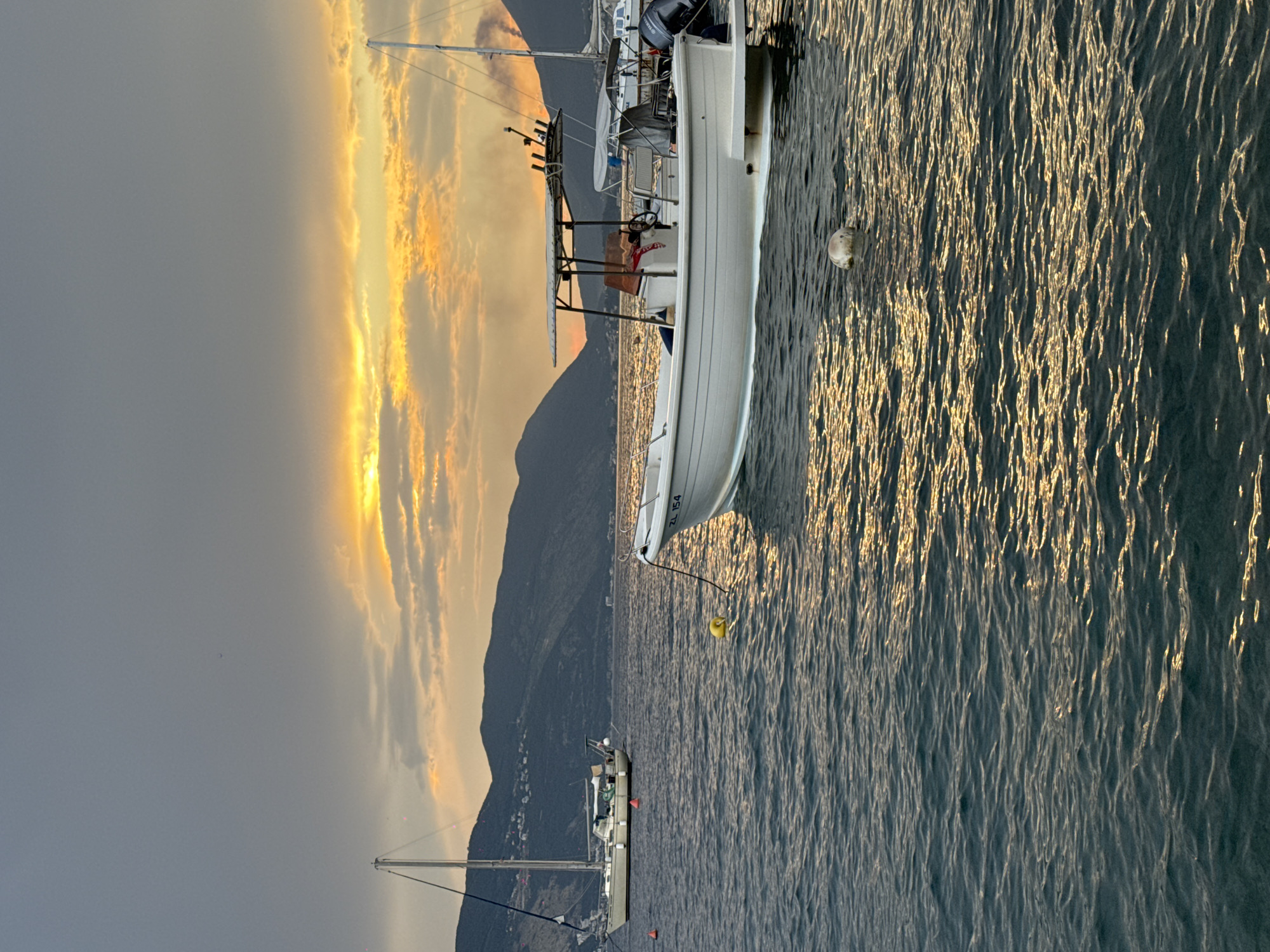 This media file captures a serene sunset scene over a body of water, likely a bay or a calm sea, with a mountainous backdrop. The sky is dominated by dramatic clouds tinged with vibrant oranges and yellows from the setting sun, contrasting with the darker, more muted tones of the mountains and the water.
Several boats are present in the scene. A white motorboat, with the visible text "ZL 154" on its side, is moored close to the viewer, its hull reflecting the golden light on the water. Another, larger sailboat is anchored further in the distance, its mast reaching towards the sky. The water itself is textured with ripples and reflections of the golden light, creating a shimmering effect.
The overall mood of the image is peaceful and tranquil, enhanced by the warm hues of the sunset and the quiet presence of the boats. The time of day is clearly sunset, indicated by the low angle of the sun and the specific colors in the sky. The weather appears calm, with the water being relatively still. The presence of mountains suggests a coastal or lakeside location, possibly in a region known for its natural beauty. No specific activities are clearly depicted, but the scene evokes a sense of stillness and the end of a day.