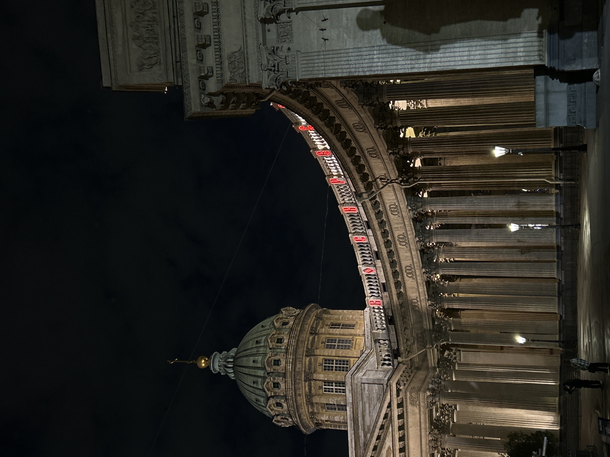 At night, the Kazan Cathedral in Saint Petersburg, Russia is depicted. The dome of the cathedral is lit with warm lighting. Above it, part of the archway is decorated with red LED signs, spanning across the frame. The archway is supported by pillars, also illuminated, that extend towards the right edge. The sky is dark. Pedestrians are visible on the right edge.