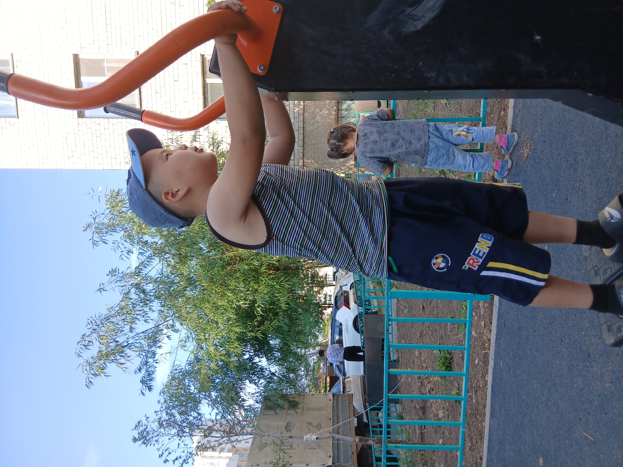 A young boy wearing a blue baseball cap and a striped tank top is standing on a piece of playground equipment in an outdoor urban setting, likely in Astana, Kazakhstan, given the context. He is gripping orange bars of the equipment with both hands and looking to his right. His dark shorts have a white and yellow stripe down the side and an emblem with the letters "BMW" and a circular logo. To the boy's right and further back, a young girl with pigtails wearing a gray t-shirt and blue jeans is also interacting with playground equipment. In the background, there is a white brick building, trees with green leaves, a light blue fence, and parked cars. A truck is also visible on the left side of the frame. The ground is paved with dark asphalt. The sky is clear and blue.