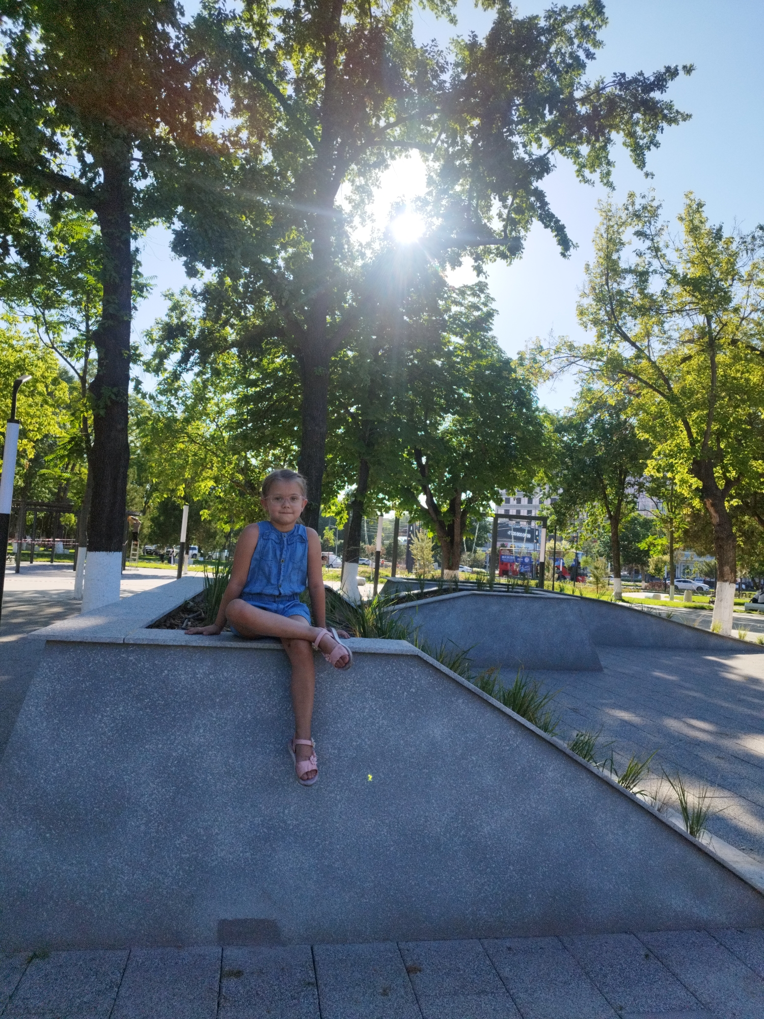 In Shymkent, Kazakhstan, a young girl with blonde hair and glasses, wearing a blue denim romper and pink sandals, sits with her legs crossed on a grey concrete ledge in a park. She is positioned to the left of the frame, facing the camera with a neutral expression. The ledge appears to be part of a skate park or similar recreational area with sloped concrete surfaces and some sparse greenery. Behind her, large trees with green foliage create a canopy, with sunlight filtering through the leaves and casting lens flare across the sky. The background shows more trees, a paved walkway, and distant buildings and streetlights, indicating an urban park setting. The lighting suggests it is a sunny day.