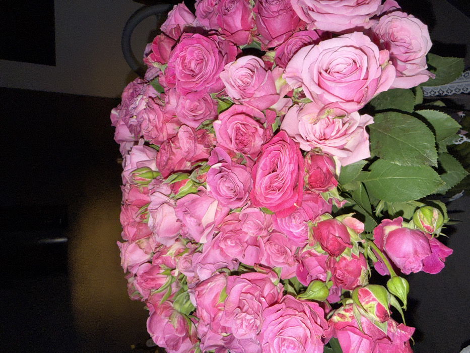 A dense floral arrangement occupies the majority of the frame, consisting exclusively of numerous pink roses and rosebuds. The roses display varying shades of pink, ranging from light pastel to vibrant fuchsia, with some petals exhibiting minor discoloration or wear. Green foliage is visible among the blossoms, particularly at the lower and left sections of the arrangement. No individuals are present. No specific actions or interactions are depicted. The background is dark and out of focus, suggesting an indoor setting with limited, ambient illumination; no architectural features or discernible objects are identifiable. The specific location within the city of Krasnoyarsk, Russia, cannot be identified from the visual data presented.