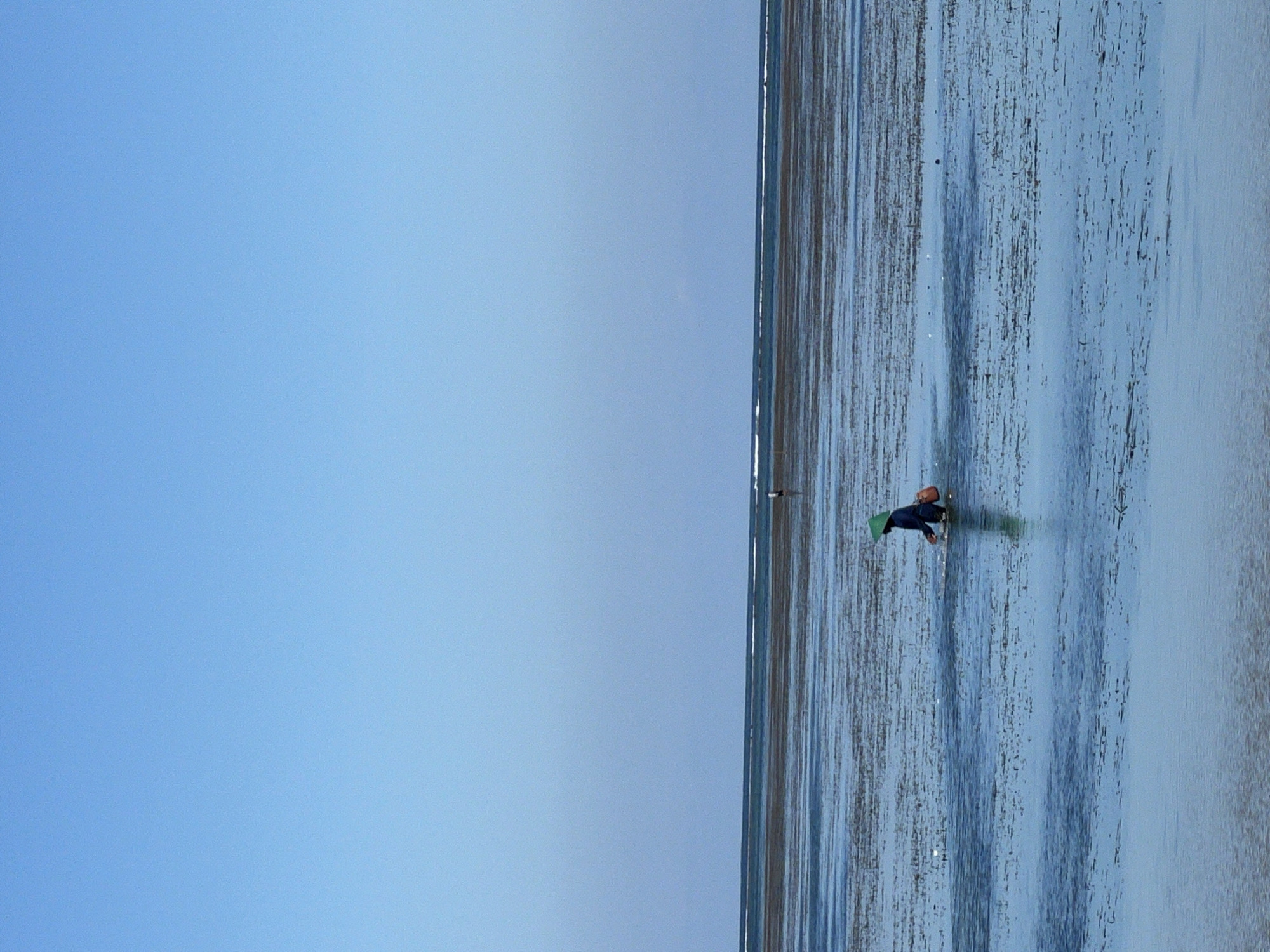 A person wearing dark clothing and a conical hat is wading in shallow water. The water is marked with dark striations, likely indicating areas of different depths or features below the surface. A narrow strip of land divides the water from a gradient-filled sky. The location is likely Denpasar, Indonesia.
