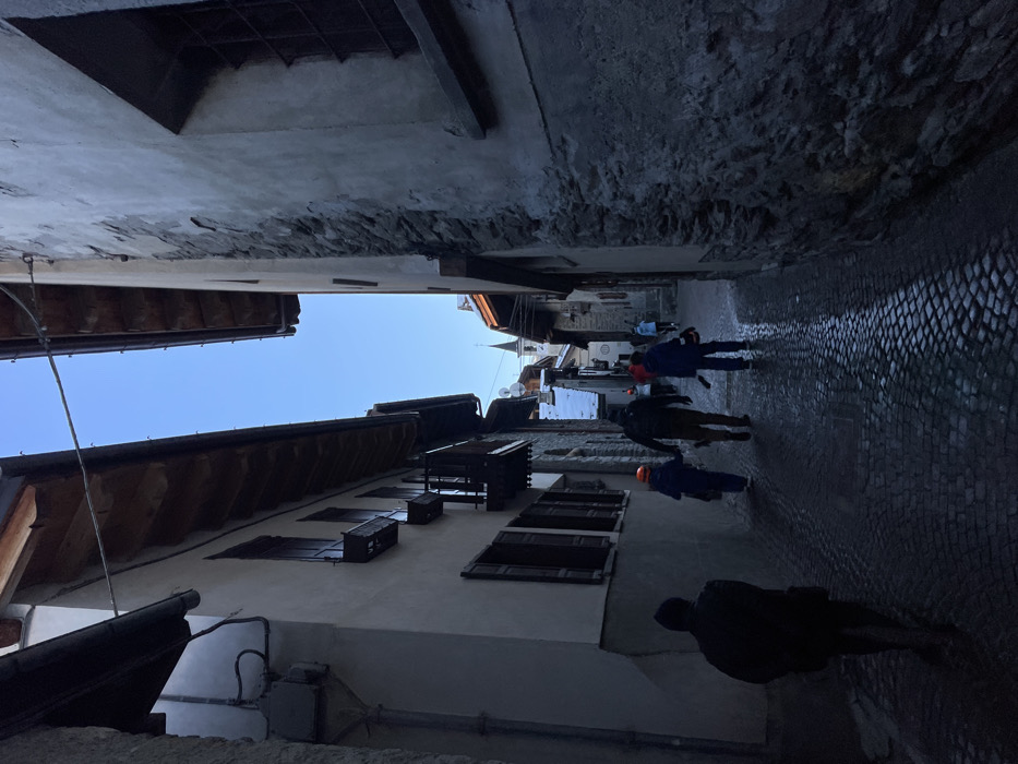 The image captures a narrow, shaded cobblestone street in Courmayeur, Italy, bordered by tall, historic buildings. The perspective is looking down the street, with pedestrians walking away from the viewer towards a distant church spire.
The street surface is paved with dark, wet cobblestones, reflecting a subtle sheen of light. Buildings line both sides; the one on the left features light-colored stucco walls with several small, dark wooden window shutters and a brown-tiled roof with visible wooden beams. The building on the right is darker, appearing to be constructed from stone or rough plaster. A sliver of clear blue sky is visible overhead, indicating daytime.
Several people are present in the scene. In the foreground, a silhouetted figure in a dark jacket walks away. Further down, two individuals are dressed in what appear to be work overalls or activewear – one in blue and orange, wearing a bright orange helmet or hard hat, and another in a dark jacket. Another person in light blue is visible deeper into the street. Their activity is simply walking, traversing the street.
The overall lighting is dim and cool, characteristic of a shaded alleyway, despite the clear blue sky. The wet ground might suggest recent rain or melting snow. There is no discernible text visible in the image.