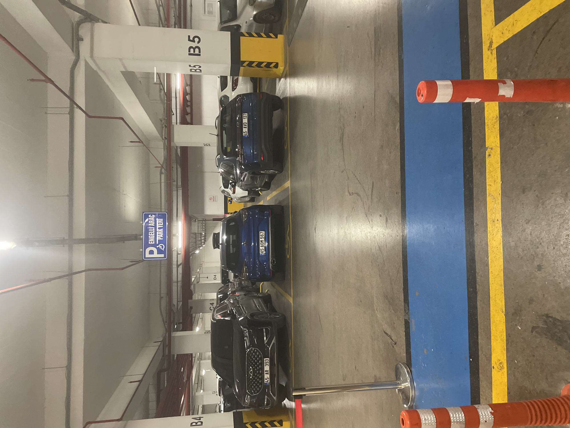 The image depicts an indoor parking garage, likely in Yunus Emre, Turkey, as indicated by the text on a car's license plate. The scene is well-lit by artificial lighting. The primary subjects are several parked cars, including a black SUV with the license plate "45 AR 393" in the foreground, a blue SUV with license plate "45 AOM 567" behind it, and a blue car with license plate "45 AVD 137" further back. The parking spaces are marked with yellow lines on the concrete floor, and there are blue and yellow stripes forming a lane.
Notable details include various signs and markings within the garage. On a white pillar, the text "B5 B5" is clearly visible. Another pillar has the text "B4" and "C4" and "D4". A blue sign with white text reads "ENGELLİ ARAÇ PARK YERİ", which translates to "Disabled Vehicle Parking Area". There are also red pipes running along the ceiling and walls, typical of fire suppression systems.
In the foreground, a red and white striped bollard is positioned on the right side of the image. The overall atmosphere is that of a functional, everyday parking facility. There are no people visible in the image, and the activity is simply cars parked in their designated spots. The time of day cannot be determined, but the artificial lighting suggests it could be day or night. The weather is not relevant as it is an indoor location.
