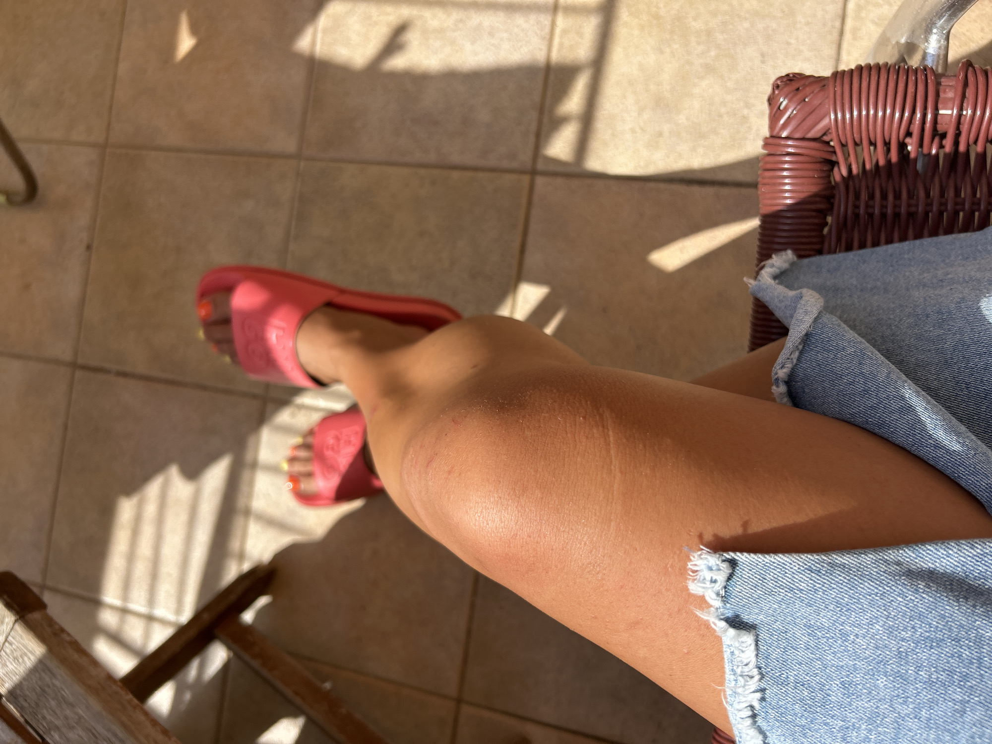 The image depicts a close-up, high-angle view of a person's legs, adorned with bright pink slides and a distressed denim skirt. The person's skin appears tanned, with visible marks on the knee. They are seated on a woven red chair, casting a shadow onto a tiled floor. Sunlight filters through what appears to be a wooden structure, creating diagonal striped shadows on the floor. The scene suggests an outdoor setting, possibly a patio or balcony, in a warm climate. The background is blurred, but the tiled floor and the chair's texture indicate a domestic or hospitality environment. The presence of a wooden chair and table in the upper left corner hints at outdoor furniture commonly found on terraces. The overall lighting and shadows suggest it is daytime, likely during a sunny period. The location is specified as Capdepera, Spain, a town known for its historic castle and coastal resorts, which often feature outdoor seating areas like the one depicted.