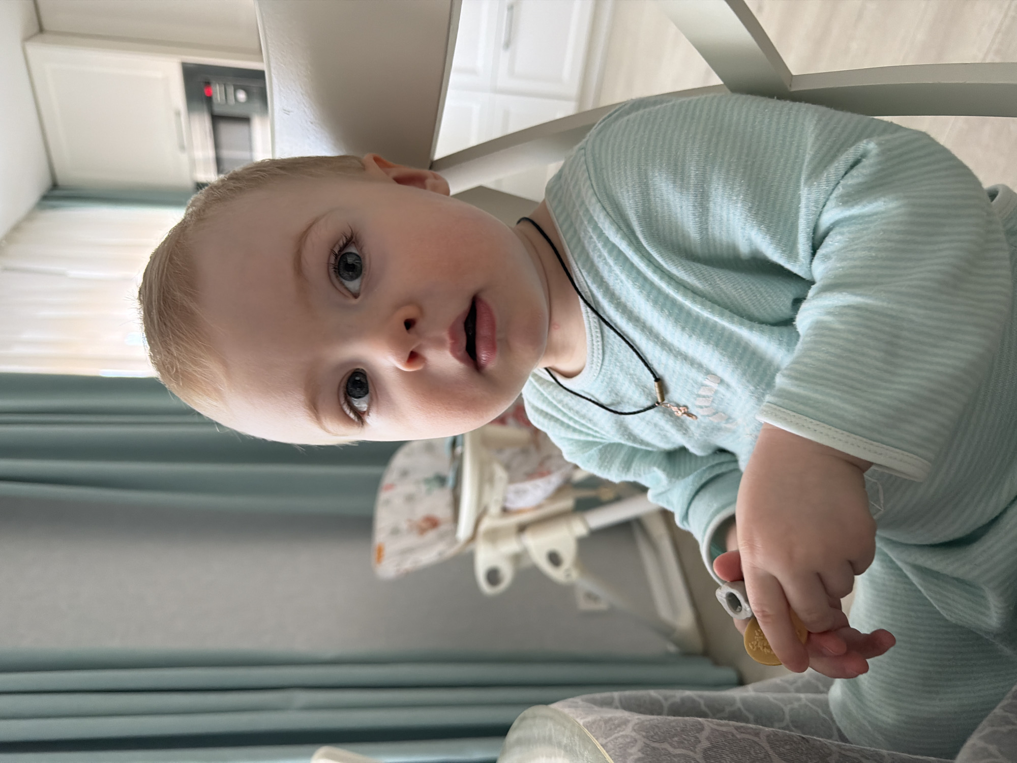 The image portrays a young child seated indoors in the city of Votkinsk, Russia. The child, appearing to be a toddler with light hair and blue eyes, is positioned facing the camera with a slightly open mouth. The child wears a light blue and white striped long-sleeved garment and a black cord necklace from which a small, metallic cross pendant hangs. In the child's right hand, a light-colored, textured teething toy is held along with a round yellow object.
To the immediate left of the child, a section of a table is visible, covered with a grey and white patterned tablecloth. The child is seated in a white chair. In the background, a beige high chair with a patterned fabric seat is partially visible to the left, positioned near light blue-green vertical window curtains. To the right in the background, a modern kitchen area is present, featuring white overhead cabinets and a built-in dark-colored microwave oven displaying red numerals. The floor throughout the visible background area is light-colored.