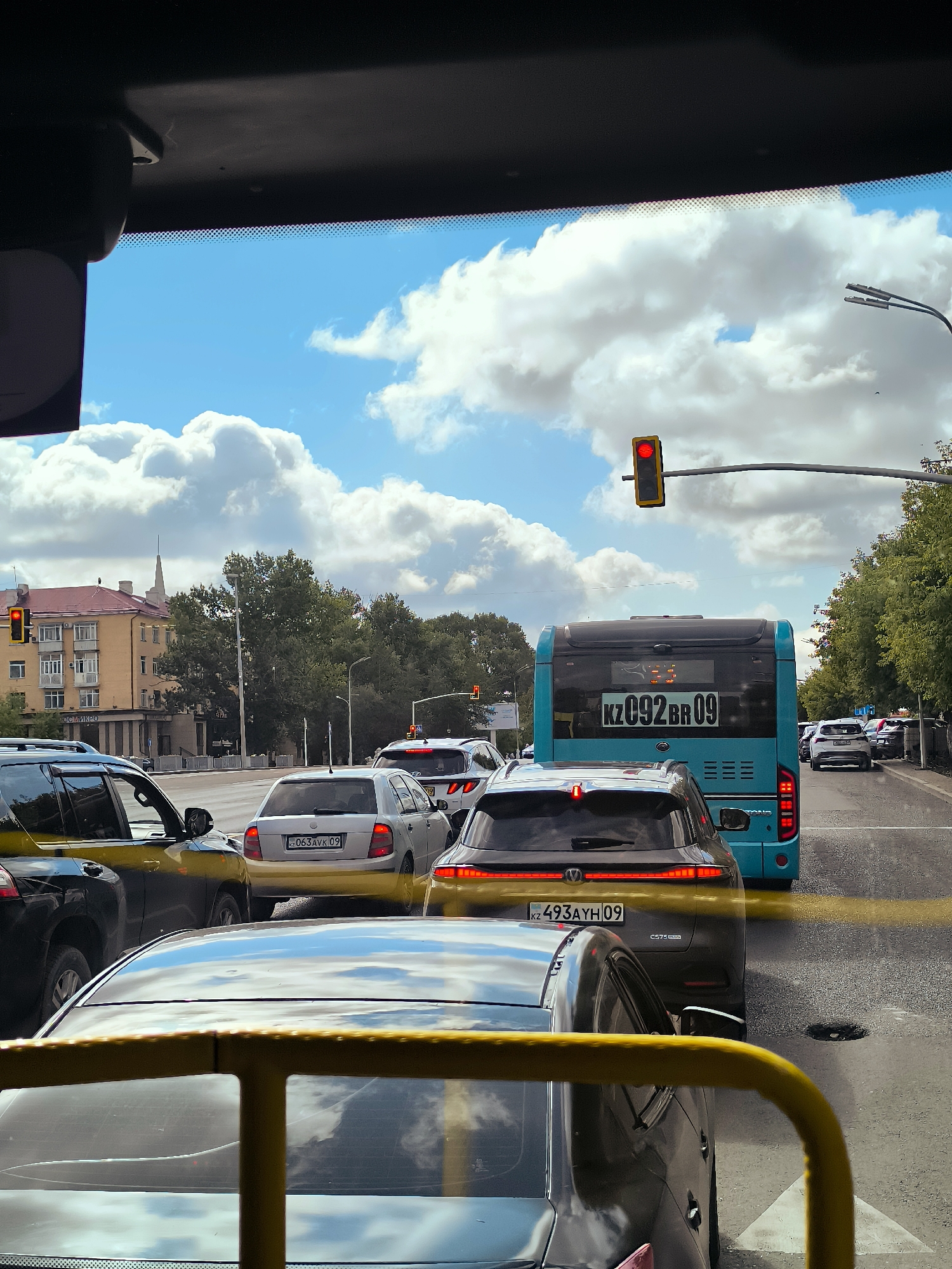 The image captures a traffic jam on a sunny day in Qarağandı city administration, Kazakhstan. A blue bus with the license plate KZ092BR09 is in the middle lane, stopped. Several cars are behind the bus, including a silver sedan with license plate 063AVK09, and a dark-colored SUV with license plate KZ493AYH09. The foreground shows the interior of a vehicle, likely a bus, with a yellow metal railing. In the background, a yellow building with a spire and trees line the street. Traffic lights are visible and appear to be red, indicating the traffic stop. The sky is blue with scattered white clouds.