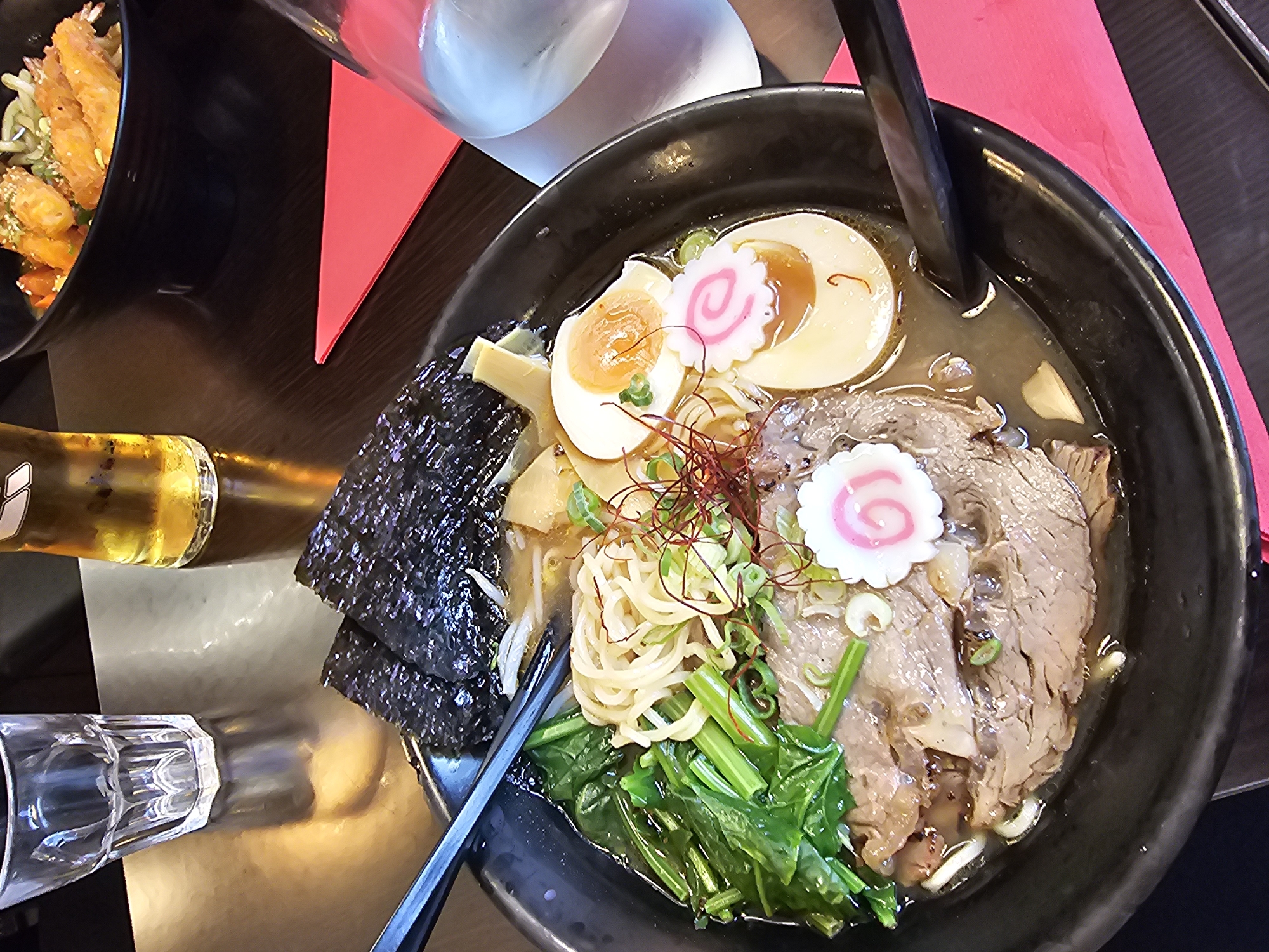 An overhead shot depicts a bowl of ramen on a dark surface, likely a restaurant table. The ramen includes broth, noodles, sliced meat, half-cut soft-boiled eggs topped with pink-swirled fish cakes, seaweed sheets, bamboo shoots, spinach, and scallions. A pair of chopsticks rests on the edge of the bowl. To the left, the neck of a glass bottle, possibly containing beer, is visible, along with an empty glass. The presence of these items suggests the image was taken in a restaurant. A red napkin is partially visible near the upper-right corner. Based on the food and possible location, the scene is possibly in Paris, France.
