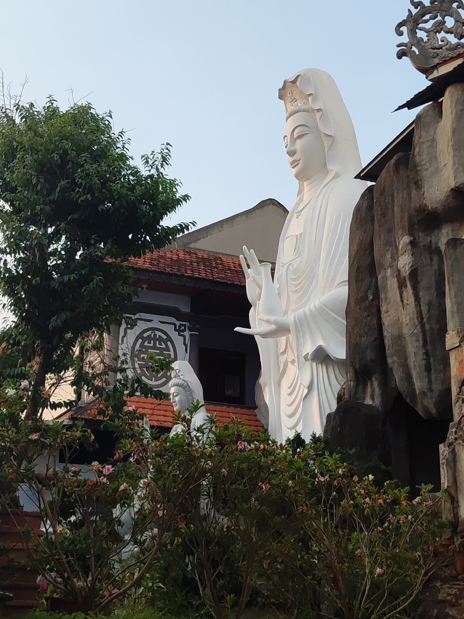A daytime shot depicts two large, white statues of Buddhist deities in Da Lat, Vietnam. The statues are surrounded by lush greenery, with trees and bushes in the foreground. Behind the statues, a building with a red-tiled roof and a circular emblem is visible. A structure resembling artificial rocks stands to the right of the statues. The sky is pale blue.