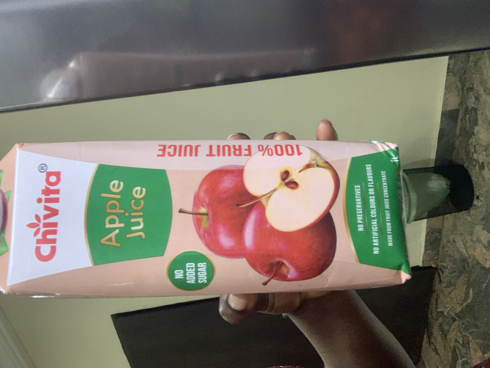 A person's hands are holding a carton of Chivita Apple Juice. The carton is predominantly light pink with a white and green design. The Chivita logo is at the top, followed by "Apple Juice" in large green lettering. A circular green label reads "NO ADDED SUGAR". Below this, there are images of red apples, one sliced in half. The side of the carton features red text that says "100% FRUIT JUICE". The bottom of the carton has a green label with white text that reads "NO PRESERVATIVES", "NO ARTIFICIAL COLOURS OR FLAVOURS", and "MADE FROM FRUIT JUICE CONCENTRATE". The carton is being held in front of a neutral-colored wall, and a dark surface, possibly a countertop, is visible at the bottom. There is a dark glass or cup placed on the countertop behind the carton. The scene appears to be indoors.