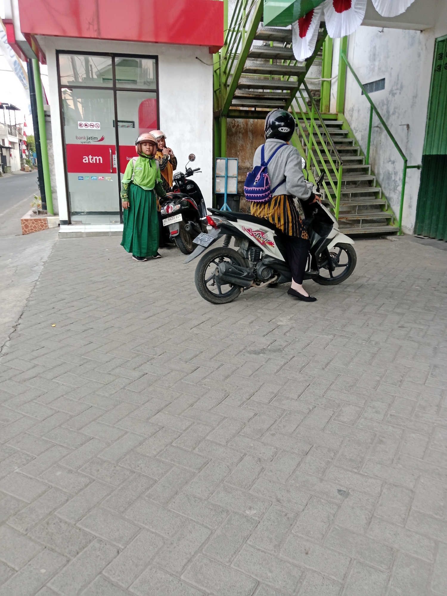 A young girl in a green hijab and dress, and an older woman in a hijab and headscarf, stand next to a black motorcycle parked in front of a Bank Jatim Syariah ATM. The ATM is housed in a small, red-and-white building with a glass facade. Behind the ATM, a green metal staircase ascends to an upper level. A woman wearing a black helmet and a grey jacket is seated on a white motorcycle in the foreground, facing away from the camera. The pavement is made of grey rectangular bricks. The scene appears to be in Kediri City, Indonesia, based on the Bank Jatim Syariah branding.