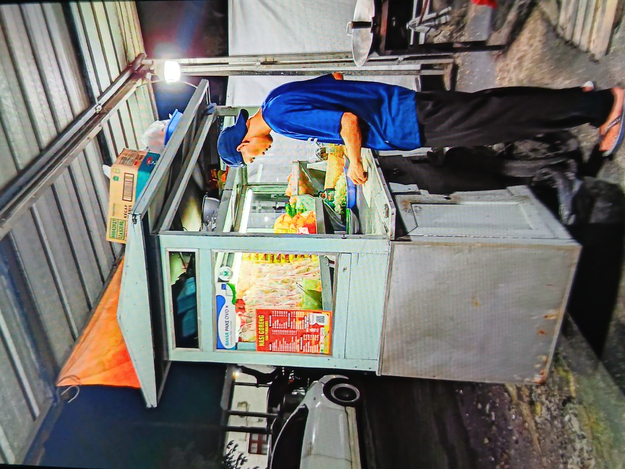 A man wearing a blue t-shirt and blue baseball cap is preparing food at a street food stall. The stall is made of metal and has a display case filled with ingredients. A menu is posted on the front of the stall, showing prices for various dishes, including "Nasi Goreng." In the background, a white car is parked, and the scene appears to be outdoors, possibly in a city like Setia Bakti, Indonesia, given the context of street food stalls. The lighting suggests it is evening or nighttime.