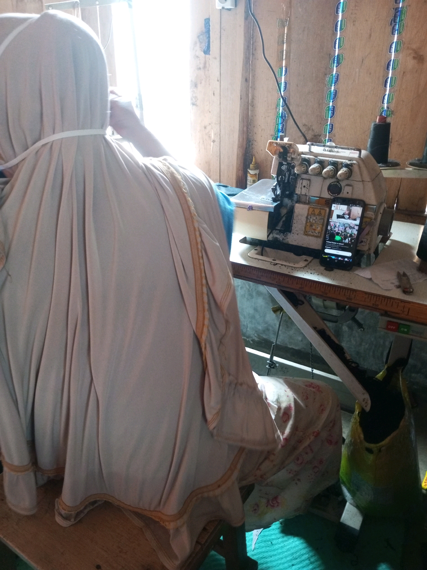 A person wearing a light-colored hijab is seated at a sewing machine in what appears to be a workshop. The person is positioned with their back to the camera, facing a sewing machine that is powered on and has a mobile phone on its surface displaying a video. The workshop is dimly lit, with natural light entering from a window on the right side of the frame. The walls are made of wooden planks, and there are various items on shelves and the workbench, including spools of thread, a bottle of liquid, and what looks like a ruler. The floor is covered with a green mat. A part of a mannequin or dummy covered in fabric with a floral pattern is visible to the right of the person. The overall scene suggests a workspace in a residential or small business setting.