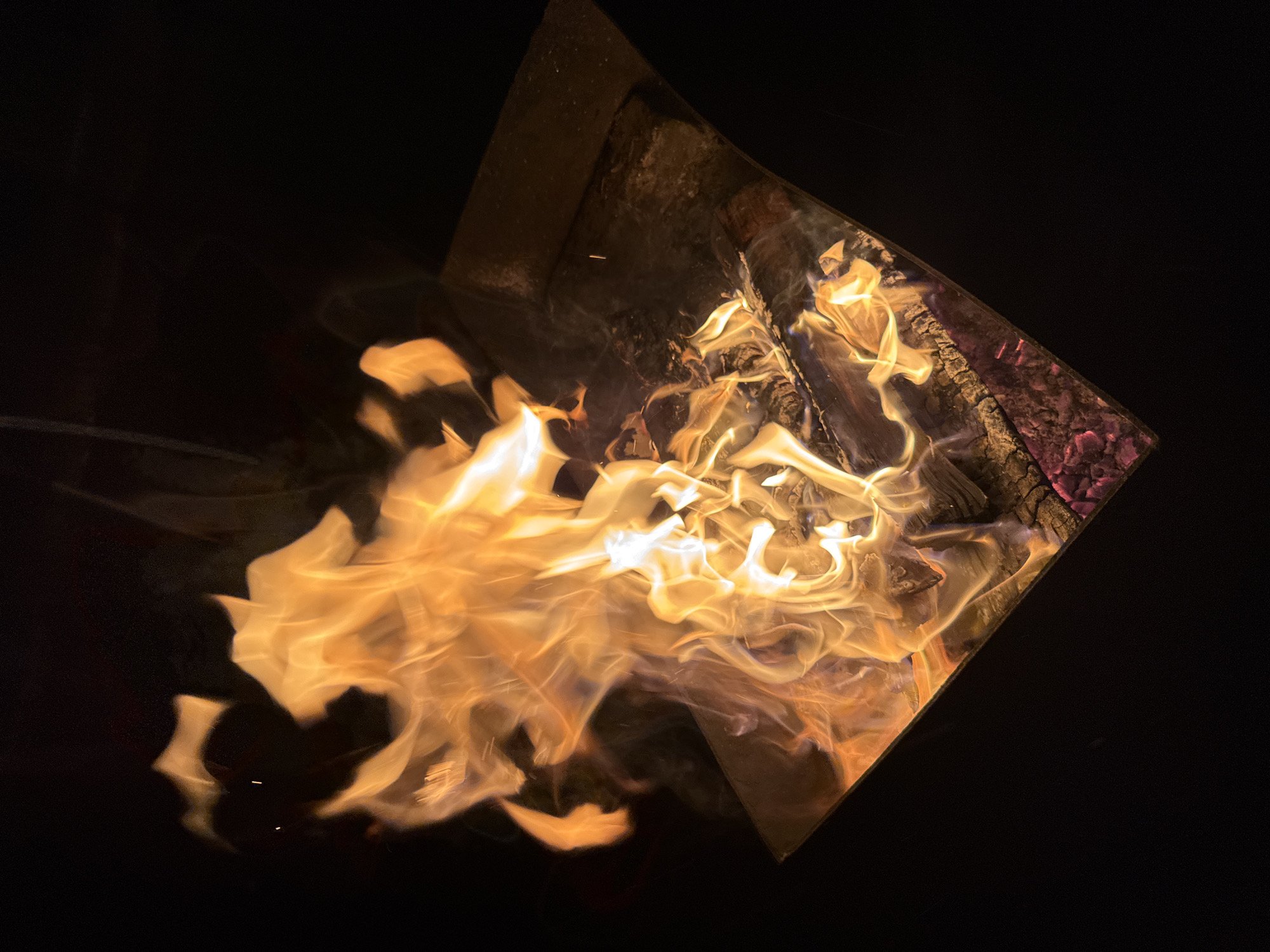 A close-up, angled shot shows a metal fire pit containing flames consuming wood. The pit's edges are visible as dark, slightly corroded metal. Inside, flames rise, with a mix of orange and white hues. Unburnt wood is visible in the background, along with dark charcoal and slight red embers. The background surrounding the fire pit is a solid, stark black. The location, Mayak, Russia, is not evident from the visual context.