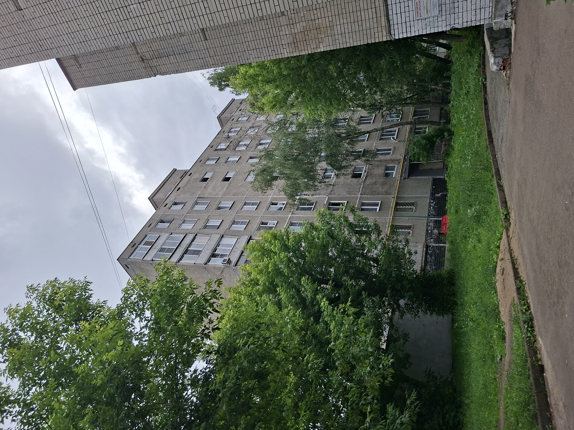 The image shows a grey, multi-story apartment building in Kazan, Russia. The building appears to be made of concrete panels, and many of the windows are visible. There are trees and grass in front of the building. To the right of the apartment building is a smaller building constructed of light-colored bricks. The sky is overcast. The scene appears to be a residential area.