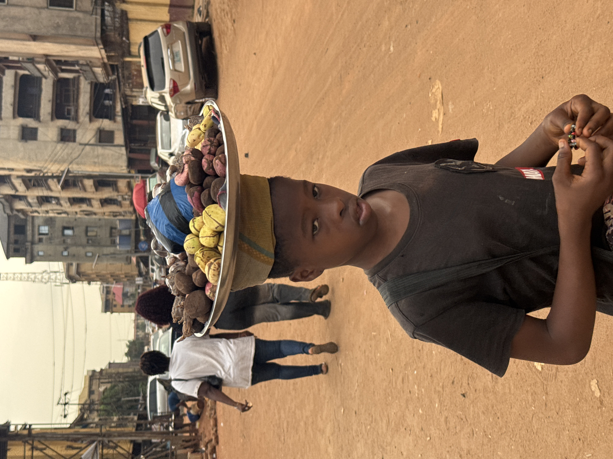A young boy in Onitsha, Nigeria, stands in a dusty street, wearing a dark shirt and carrying a tray piled high with fruits and vegetables on his head. He holds a small beaded object in his hands, appearing to examine it. Two women walk behind him, along the same street. Cars are parked along the side of the street, and several multi-story buildings are visible in the background, suggesting a busy urban environment. The air has a somewhat hazy appearance.