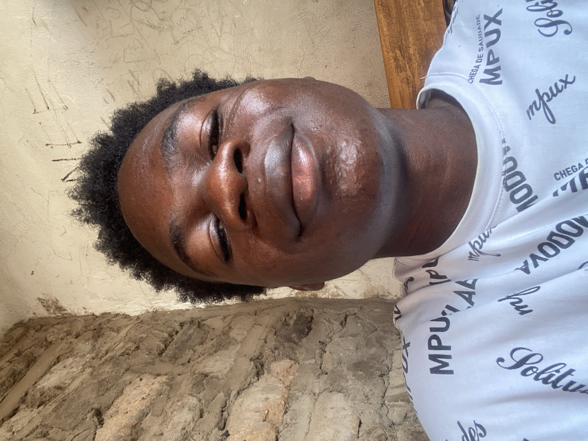 A close-up, slightly angled portrait of a dark-skinned young man is presented against a textured background. The man's eyes are nearly closed, suggesting a relaxed or introspective mood. He has short, tightly curled hair and full lips. He is wearing a white t-shirt printed with black lettering, partially obscured but containing the word "MPUX".
The background is divided into two distinct parts. The upper section is a pale, textured wall with faint markings. The lower section appears to be a rough, unfinished wall with exposed concrete or stone.
While the question mentions Mararaba, Nigeria, the image itself contains no elements to definitively confirm this location. Therefore, the location cannot be determined with certainty from the image alone.