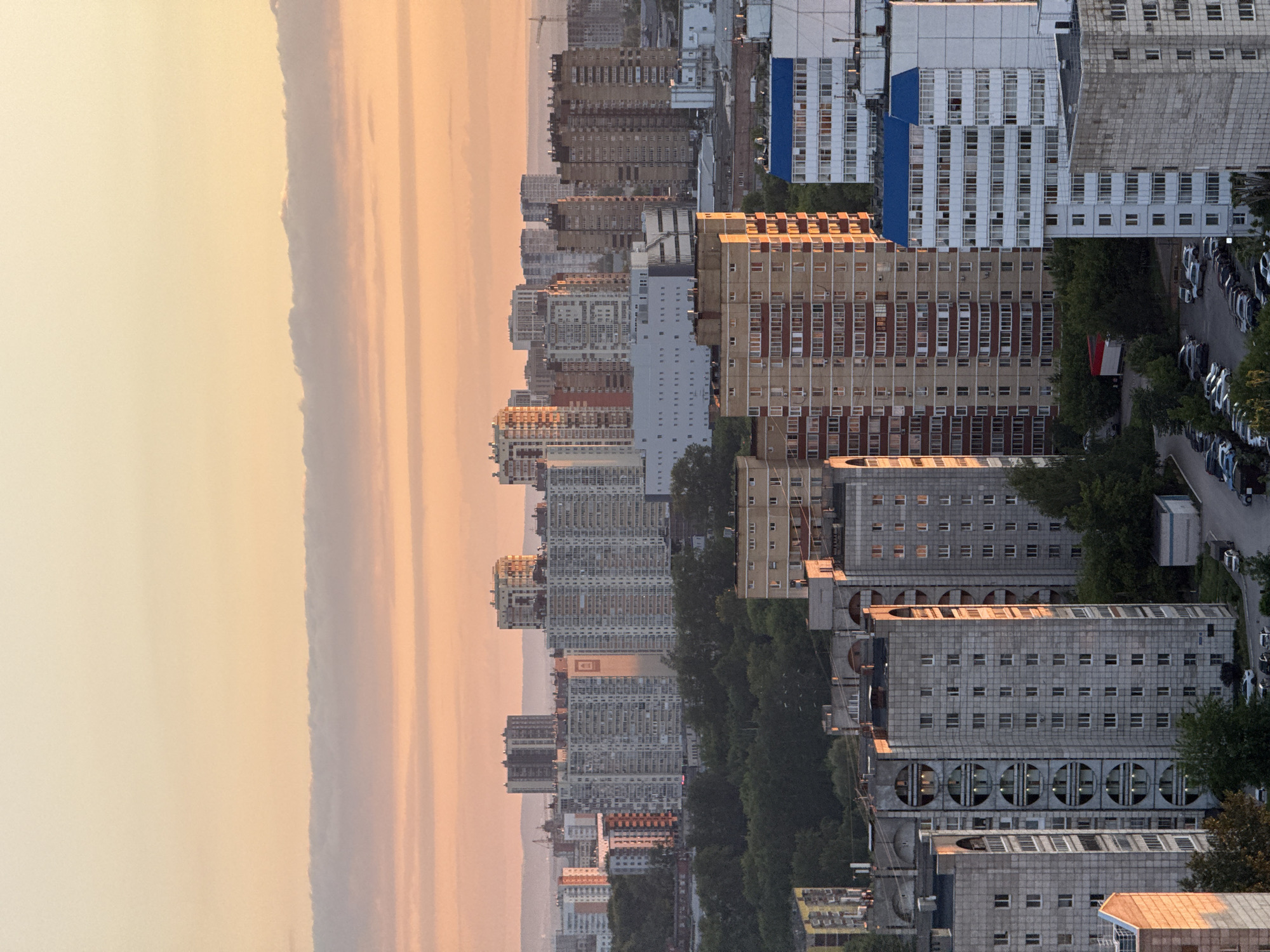 This panoramic image captures a dense urban landscape in Dikaya Gar', Russia, bathed in the soft, warm light of either dawn or dusk. The sky, occupying the upper portion of the image after correct orientation, displays a gentle gradient of colors, transitioning from a pale yellow-orange at the top to a muted, hazy pink-orange closer to the horizon, suggesting the presence of atmospheric haze or distant clouds.
The city is characterized by numerous multi-story residential buildings that stretch across the midground and foreground. These structures exhibit a variety of architectural styles, mostly functional, with facades ranging from light grey and white to beige, brown, and red. Many buildings feature repetitive window patterns, and some in the foreground display distinct architectural elements, such as large circular openings on their lower levels. The buildings are densely packed, with rows of dark green trees lining streets and interspersed throughout the urban fabric, providing a contrast to the concrete structures.
In the lower right section of the image (foreground right, after rotation), a street is visible with numerous parked cars, predominantly light-colored. A faint silhouette of distant hills or mountains can be seen along the horizon line, separating the cityscape from the sky. There are no visible people or discernible text on any of the buildings or signs. The scene conveys a sense of quiet urban vastness illuminated by the serene, diffused light of the early morning or late evening.