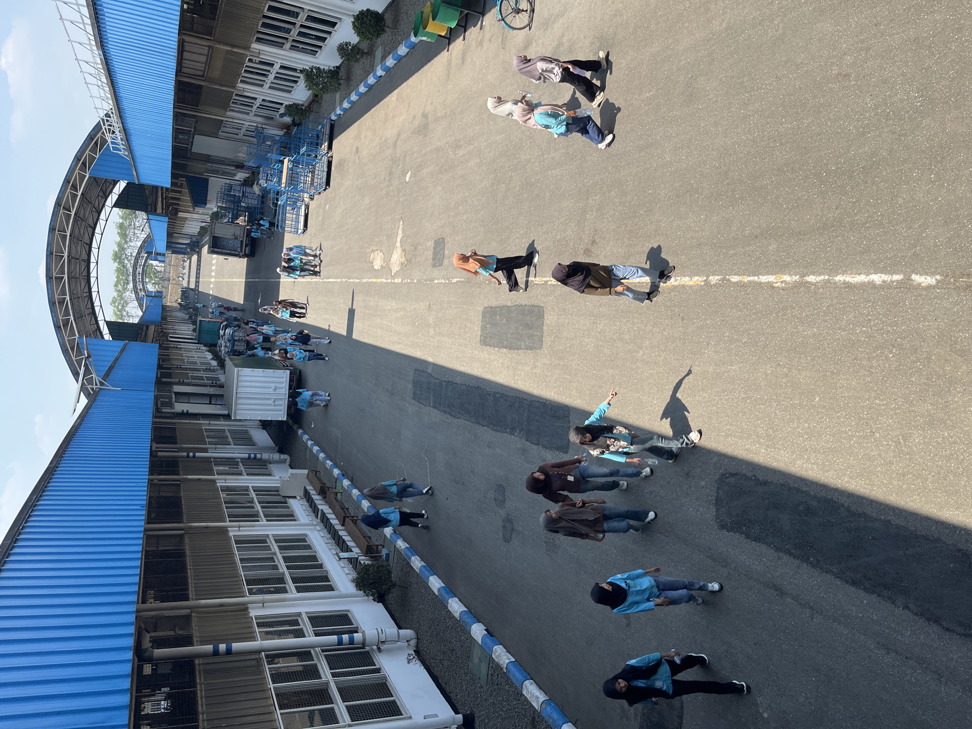 The image depicts a view from above of an industrial area, likely a factory or warehouse complex, under a clear sky. The architecture features long, blue-roofed buildings with white facades and numerous windows. An asphalt pathway runs between the buildings, populated by several individuals, mostly women wearing hijabs and casual clothing. Some are walking alone, while others are in small groups, engaged in conversation or movement. Several light-colored trucks and blue metal carts are visible, suggesting ongoing logistical activities. The ground is paved with asphalt, marked with white lines. The presence of trees and lush vegetation along the edges of the pathway indicates an outdoor setting within an industrial zone. The scene appears to be a normal workday, with people moving about their business in a sunlit environment. The location is identified as Pelang, Indonesia, though this specific detail is not visually verifiable within the image itself.