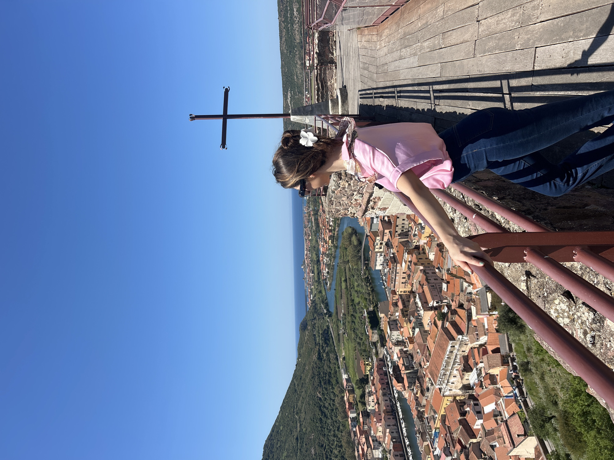 A vertical shot shows a young woman, probably in her late teens or early twenties, peering over the edge of the Castello di Bosa in Sardinia, Italy. She is clad in dark blue jeans, a light pink shirt, and sunglasses. A white flower is tucked into her dark brown hair. She appears to be Caucasian, given the pale skin tone of her hand visible as she clutches a rusted railing. In the background, the city of Bosa spills out below with its distinctive multi-colored rooftops and a narrow river meandering through the town. In the top left of the frame, a dark metal cross stands silhouetted against a cloudless, azure sky. The wooden platform she's standing on stretches across the top of the image, creating depth. The photograph is taken in bright daylight, casting stark shadows that emphasize the textures and materials of the structures and landscapes in view.