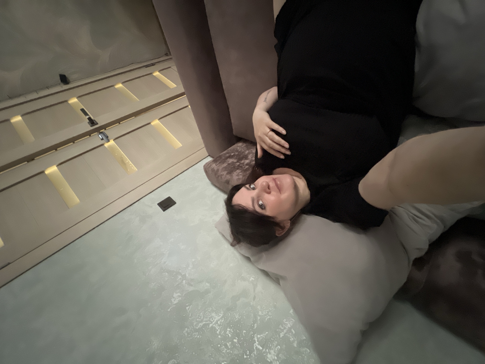 A woman lies on a light gray pillow on a textured light blue floor. She is wearing a black garment. Her left hand, bearing a ring, rests near her face. Part of a brown couch is visible behind her. Wardrobe doors with gold accents are on the left of the image. The location is identified as Noviy, Russia.