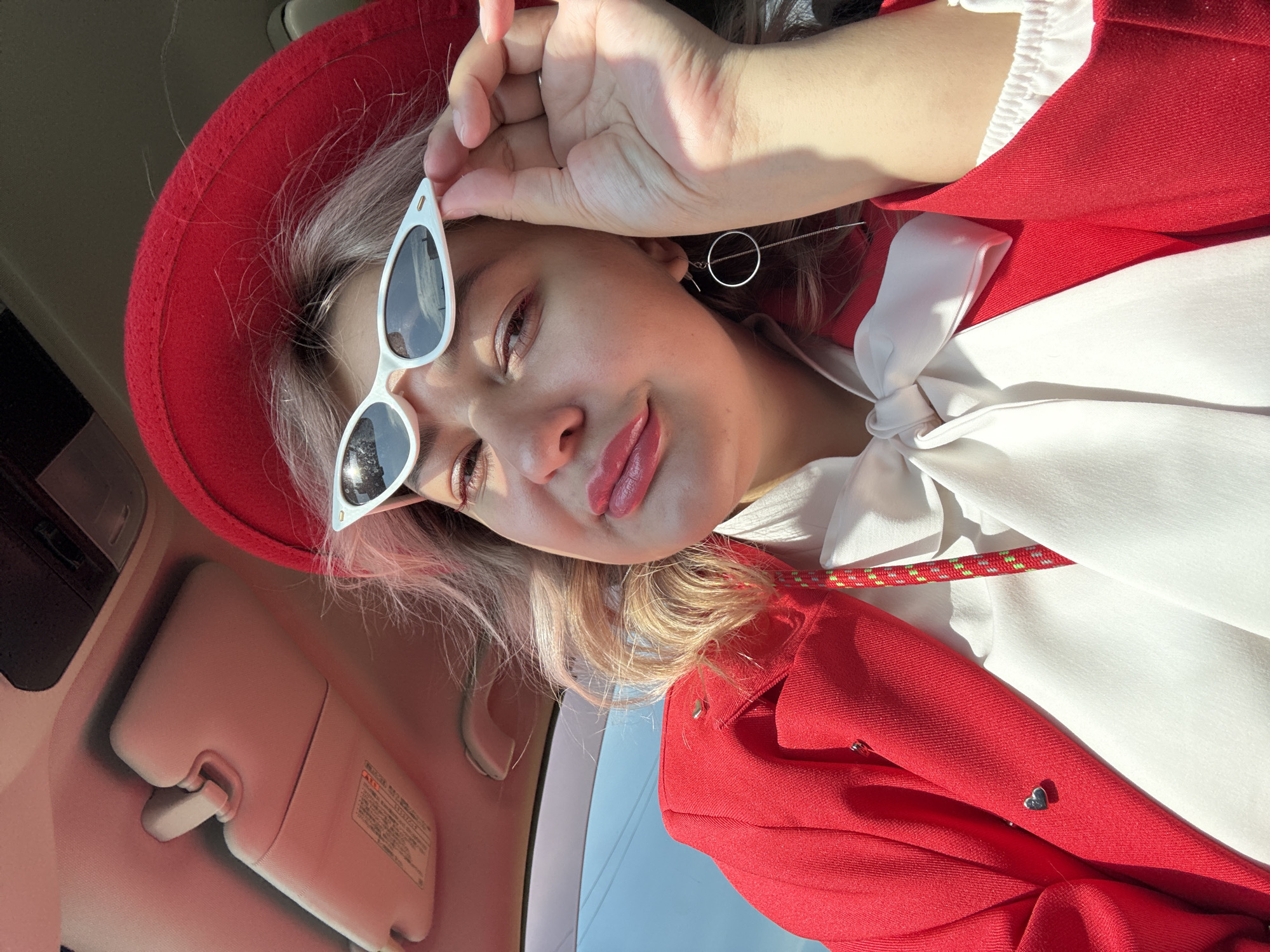 A woman with blonde hair and a red hat is inside a car. She is wearing a red jacket, a white blouse with a large bow, white cat-eye sunglasses, and long silver earrings. Her right hand is raised, holding her sunglasses. The sun is shining, casting shadows inside the car.