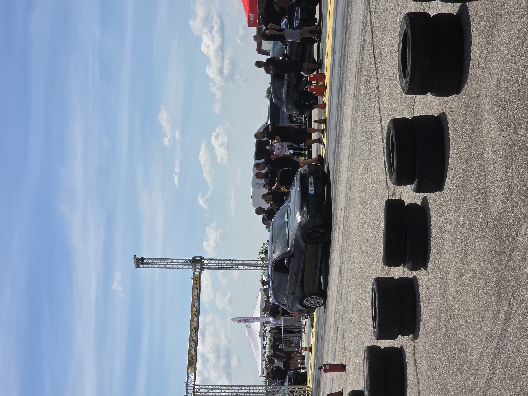 This image depicts a drag racing event at the Akatsievo airfield in Akatsievo, Bulgaria. The scene is set on a paved asphalt surface, likely a runway or dedicated drag strip, marked with yellow lines. In the foreground, several stacks of black tires are visible, serving as visual markers or perhaps part of the event's setup.
A black BMW station wagon is positioned on the track, appearing ready for a race. Several individuals, men and women, are present, some standing near the car and others gathered in the background, suggesting spectators or event staff. Tents and a red canopy are visible further back, indicating support structures for the event. A metal gantry with electronic equipment, possibly a timing system, spans across the track in the mid-ground, with a sign overhead that reads "BG Drag Team." The sky is a bright blue with scattered white clouds, indicative of fair weather. A fire extinguisher in red is placed on the track near the start line.