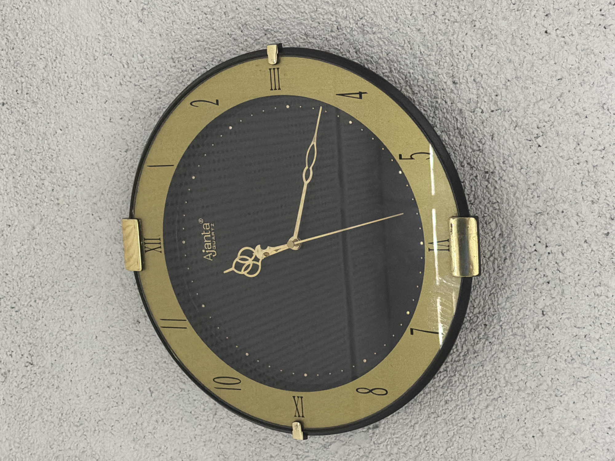 A round wall clock featuring a prominent gold-colored outer ring and a dark gray inner face is centrally mounted on a light gray, textured wall. The inner face of the clock has a subtle grid-like pattern and is adorned with small gold dots marking the minutes. The hours are indicated by a mix of Roman and Arabic numerals: XII, III, VI, and IX are presented in Roman numerals, while 2, 4, 5, 7, 8, and 10 are shown in Arabic numerals. A simple vertical line marks the 1 o'clock position.
Three gold-colored hands — an hour hand, a minute hand, and a second hand — protrude from the center. The hour and minute hands both feature an elaborate, looped design near their base. The clock displays the time as approximately 1:07:25, with the hour hand just past the 1, the minute hand pointing to the 7-minute mark, and the second hand at 25 seconds.
Four small, rectangular gold-colored decorative elements are attached to the outer edge of the clock at the 12, 3, 6, and 9 o'clock positions. The brand name "Ajanta" and the word "QUARTZ" are visible in white lettering on the upper left quadrant of the clock face. The background wall exhibits a rough, granular texture, possibly concrete or stucco, and is evenly illuminated. A slight reflection is visible on the clock's glass cover.