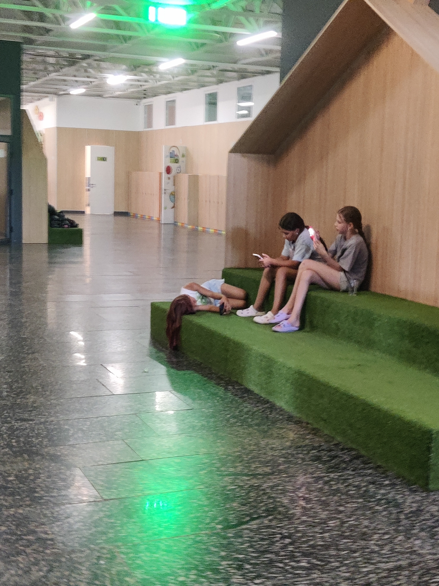 The image depicts an indoor scene, possibly a school or community center hallway, with a reflective tiled floor. In the foreground, a young girl with long brown hair is lying on a green, tiered seating area, appearing to be relaxed or sleeping. Further back, two other young girls are seated on the same green seating structure, each looking at their mobile phones. The girl on the left is wearing a light-colored shirt and shorts, while the girl on the right wears a grey t-shirt and shorts. A glass is visible next to the girl on the right. The hallway features wooden wall panels and white doors, and fluorescent lights are visible on the ceiling, one of which emits a green glow. The environment suggests a modern, clean, and somewhat minimalist design. There is no visible indication of the image being located in Kyiv, Ukraine.