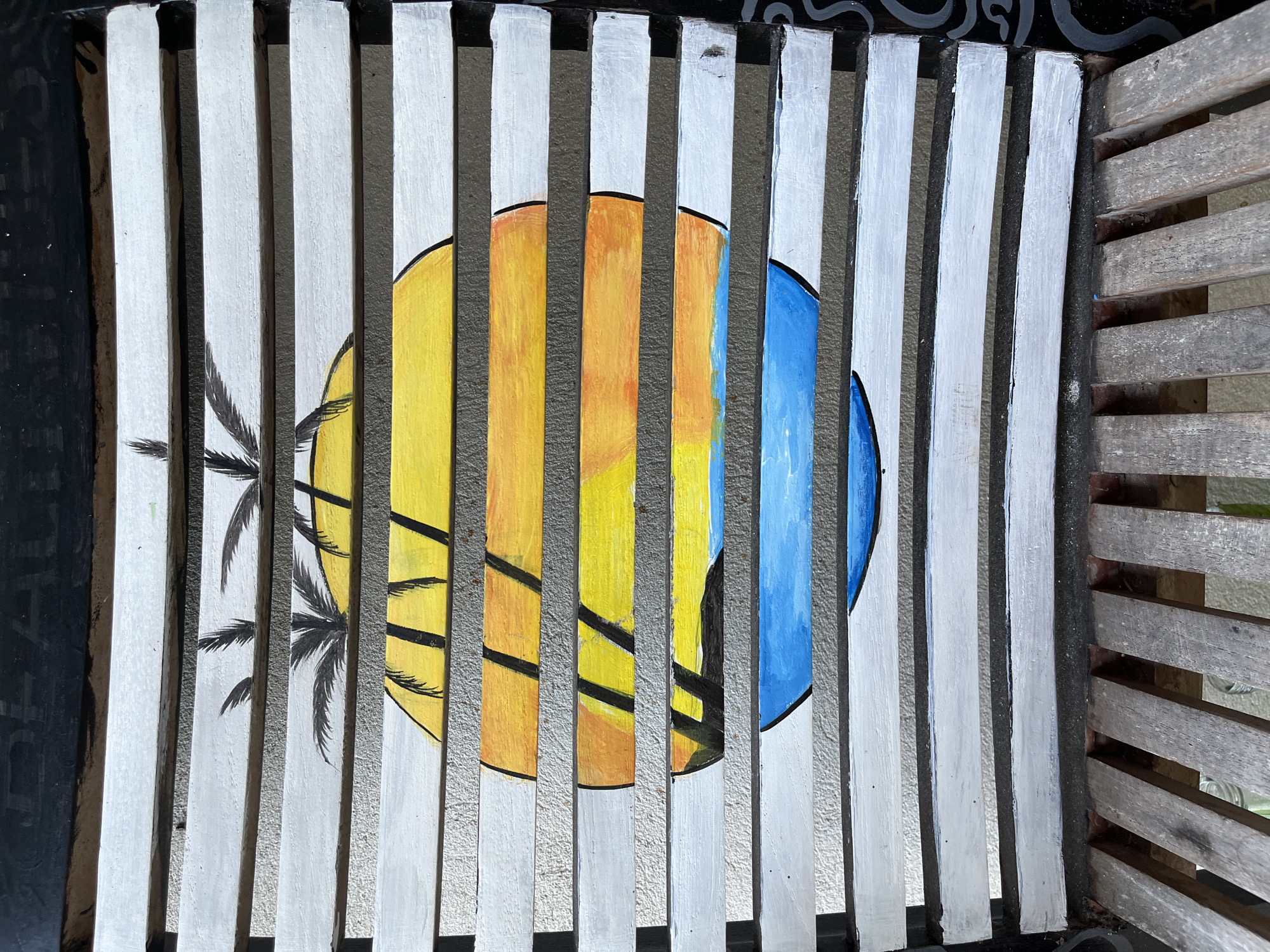 This image shows a close-up, overhead view of a wooden chair that has been painted with a sunset scene. The chair's slats are painted white, and the artwork is painted across them. The scene depicts a large, segmented circle representing the sun, with bands of yellow, orange, and blue painted within. Silhouetted palm fronds are painted above the sun. The chair is located on a surface that appears to be concrete. The specific location in Legonipah, Indonesia is not visible in the image.