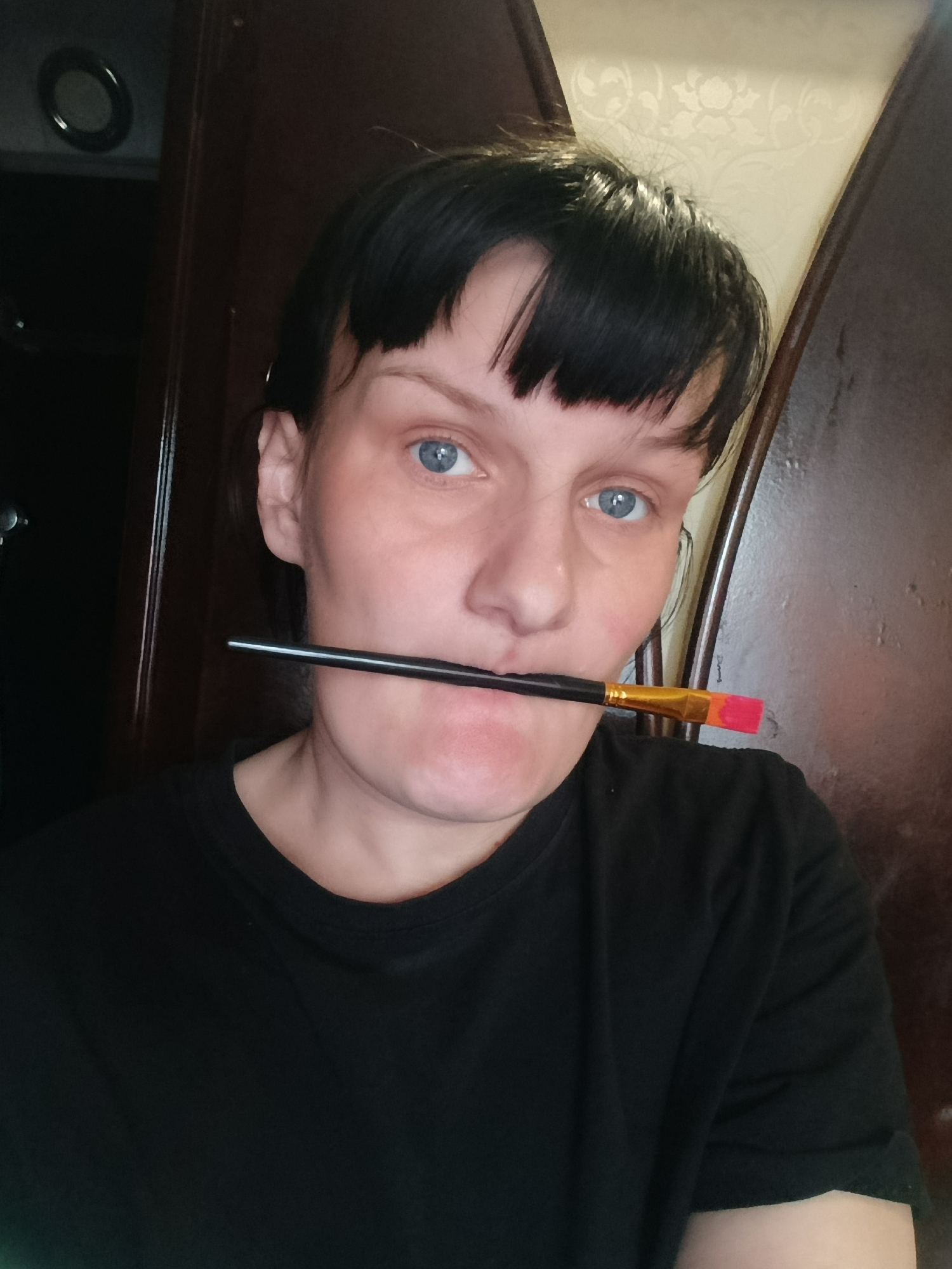 A close-up, chest-up portrait features a person with short, dark hair and bangs, and striking blue eyes, looking directly at the viewer. She holds a black-handled paintbrush horizontally in her mouth; the brush end, visible on the right, is tipped with vibrant red and orange colors. She wears a plain black t-shirt. The person's expression is neutral to slightly serious.
The background consists primarily of dark brown, possibly wooden, wall or door panels. To the upper left, a dark circular object with a reflective rim is visible, which could be a light fixture or vent. A narrow strip of lighter-colored wallpaper with a subtle floral pattern is visible in the upper right background. The subdued indoor lighting suggests the photograph was taken in an interior setting, possibly in the evening or a room with dim ambient light. There is no visible text in the image. The location context for this image is Foki, Russia.