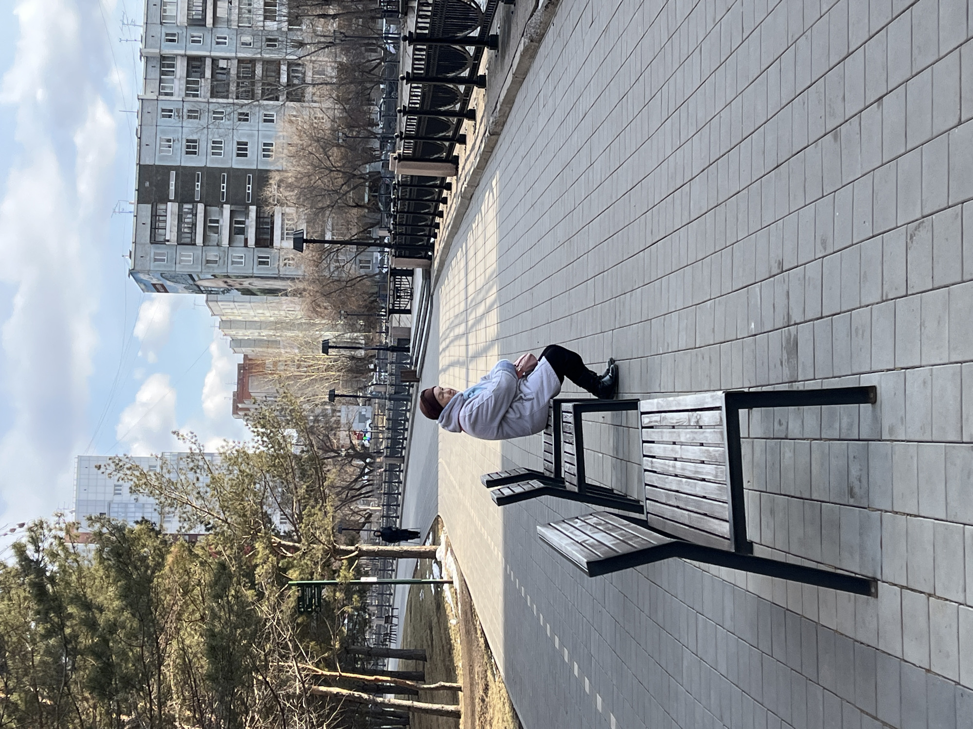 A person is sitting on a bench in a paved area in Krasnoyarsk, Russia. The person is wearing a light-colored coat and dark pants. The paved area has multiple benches with black frames and wooden slats. Buildings are visible in the background, including an apartment building. Trees without leaves are also in the background. A line of black metal fencing is present along the pathway. The weather appears to be sunny. The area is a park or public space.