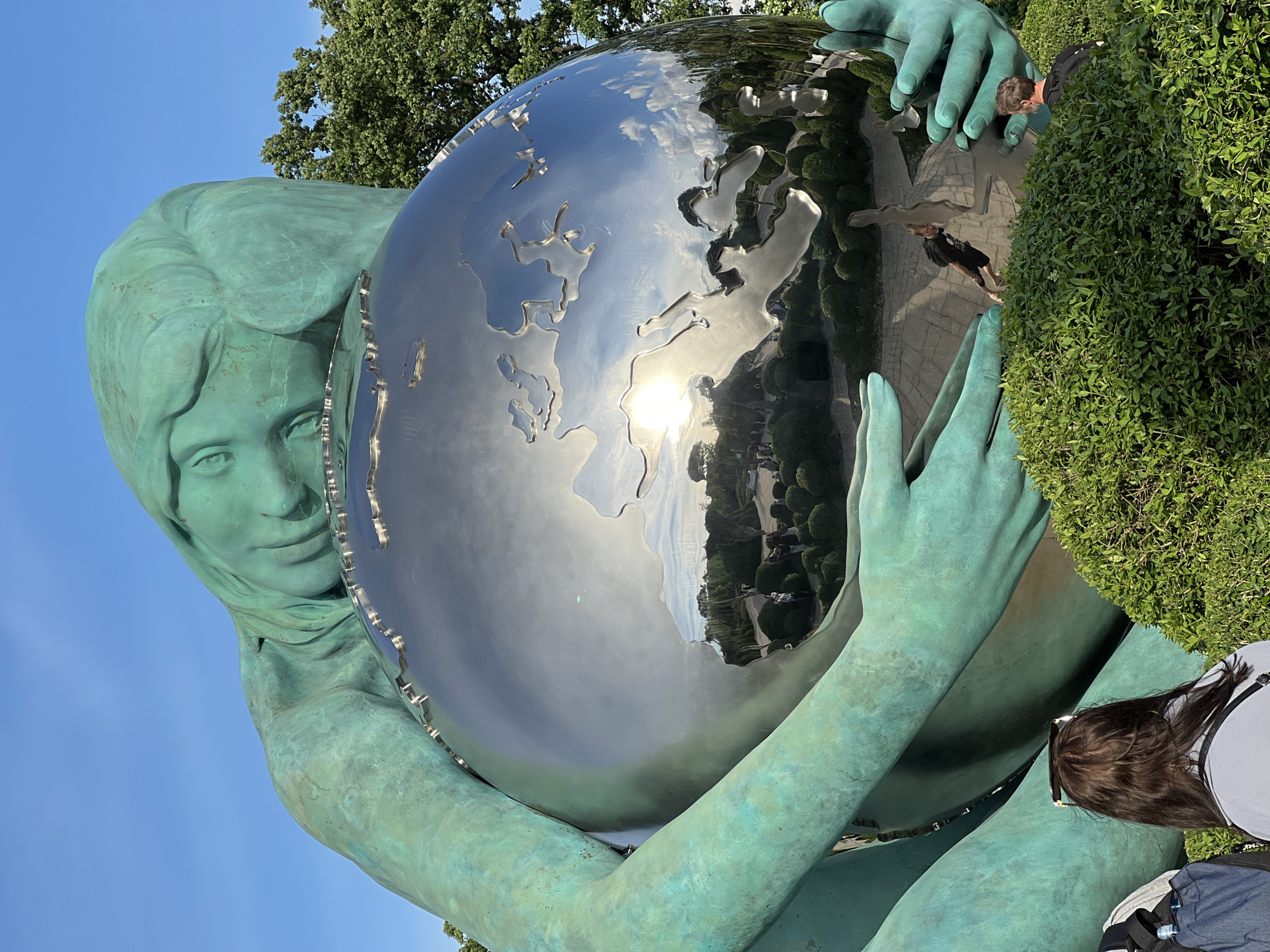 A statue of a woman with greenish-blue skin is shown in the image. She is holding a shiny metal globe with cutouts of continents in her arms. The globe reflects the surrounding environment. A pathway and manicured green bushes are visible in the reflection, along with some people. The sky is clear and blue. A tree is visible in the background above the statue's head. The bottom right corner of the image shows the back of a person's head with long brown hair. This statue is located in Krasnodar, Russia.