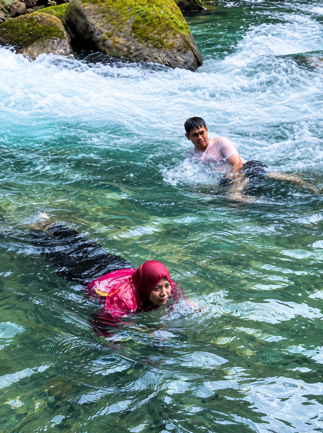 In the clear, green waters of the Cipunaga River in Gunung Ambat, Indonesia, two individuals are engaged in swimming. The image captures a man in a pink t-shirt and black shorts, actively swimming in the upper portion of the frame. In the foreground, a woman wearing a red hijab and a pink shirt is swimming towards the viewer. The river flows over a rocky bed, with large moss-covered rocks visible in the background. The water is turbulent in places, indicating a moderate current.