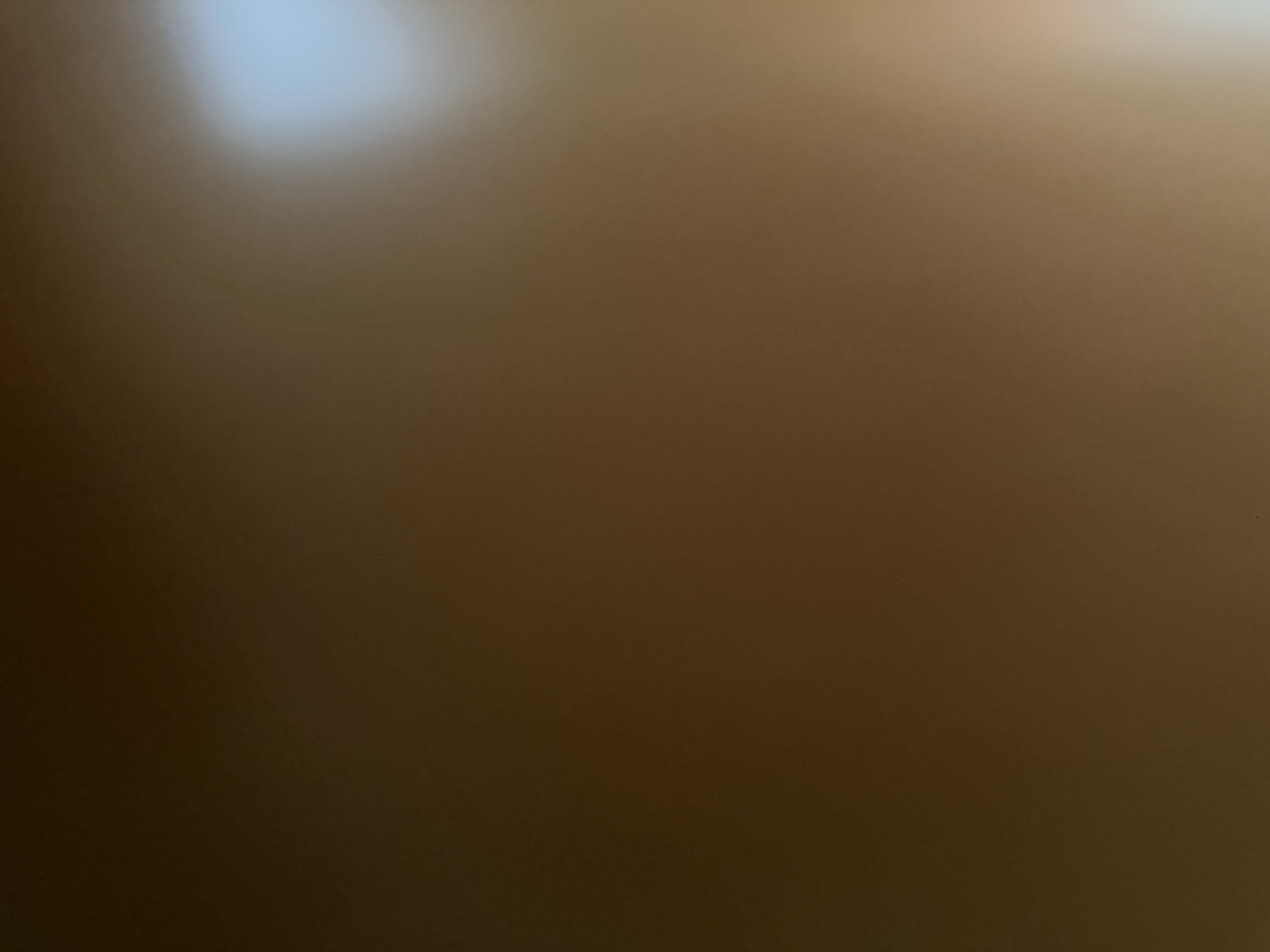 The image shows a blurred, abstract view of what appears to be a wall or a large flat surface. There is a bright light source in the upper right quadrant, casting a soft glow and creating a gradient of light and shadow across the surface. The overall color palette is dominated by shades of brown and tan, with the bright light area appearing almost white or light blue. There are no discernible people, objects, or specific environmental features visible to indicate a location within Belgrade, Serbia, or any other urban setting. The image is entirely abstract and lacks any identifying details.