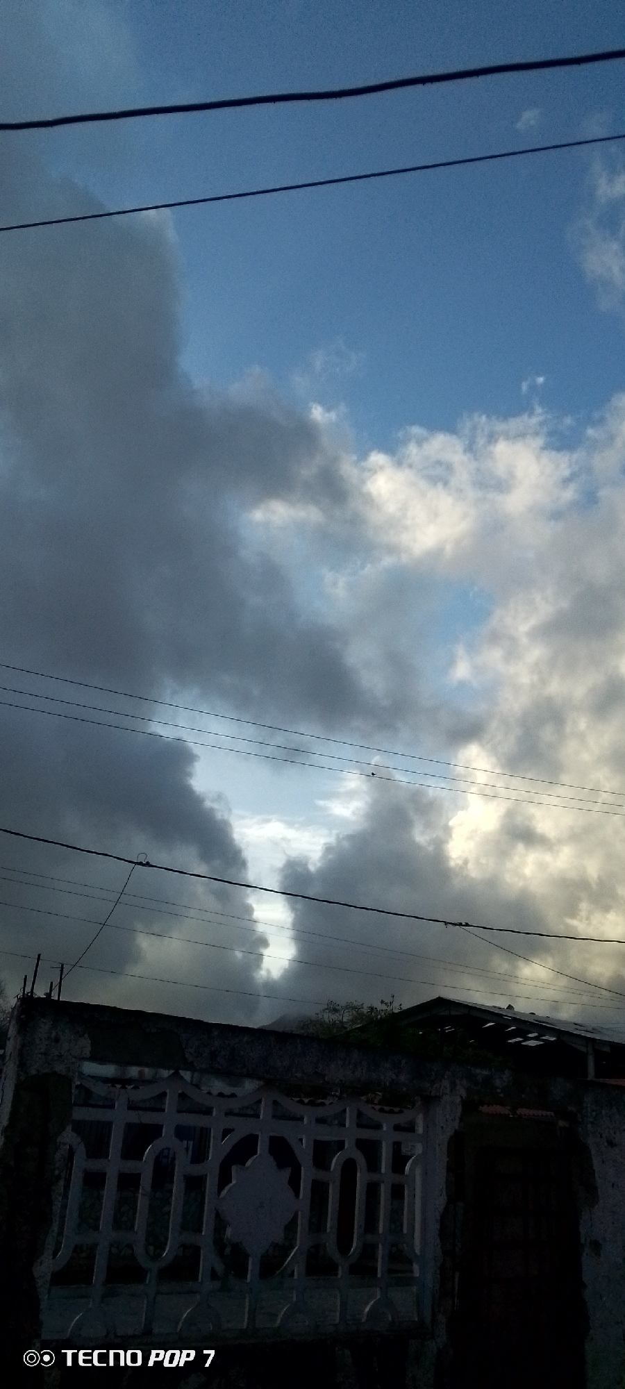 The image shows a cloudy sky with some patches of blue. There are several power lines crisscrossing the frame, and a low-lying structure, possibly a wall or a building, occupies the bottom portion of the image. The structure appears weathered and has a decorative grate-like opening. The overall scene suggests an outdoor environment, possibly a residential area, in Altagracia, Venezuela. There are no people or specific actions depicted. The lighting indicates it might be late afternoon or early evening.
