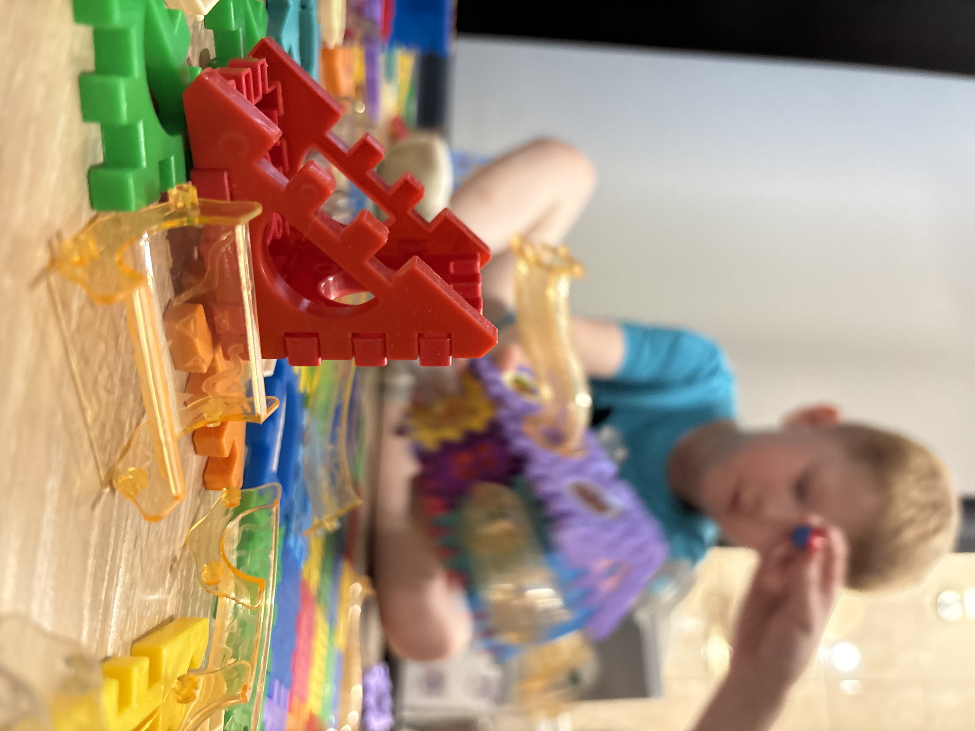 In this close-up, low-angle shot of a playroom floor, a young boy with blond hair sits in the blurred background, wearing a blue t-shirt. He is focused on a colorful toy structure, a purple castle-like piece, which he is manipulating with his hands. Another person's hand, partially visible, is placing a small red and blue object near the boy's face, suggesting they are playing together or assisting him. The foreground is dominated by an assortment of building toy pieces in vibrant colors, including red, green, orange, and yellow, arranged on a wooden floor. Some of these pieces are transparent orange plastic connectors and bright red interlocking blocks, hinting at a construction or marble run set. The background shows a hint of a wall and flooring, typical of an indoor domestic environment. The image does not provide any information about the specific location within the city of Izhevsk, Russia.