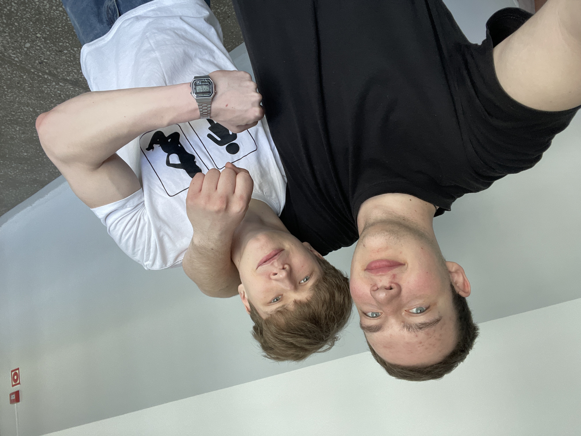 Two young men are posing for a selfie indoors. The man on the left is wearing a white t-shirt with a logo and a silver digital watch. The man on the right is wearing a black t-shirt. Part of the background shows a gray floor, a white wall, and a fire alarm box on the wall. The city location is Severodvinsk, Russia.