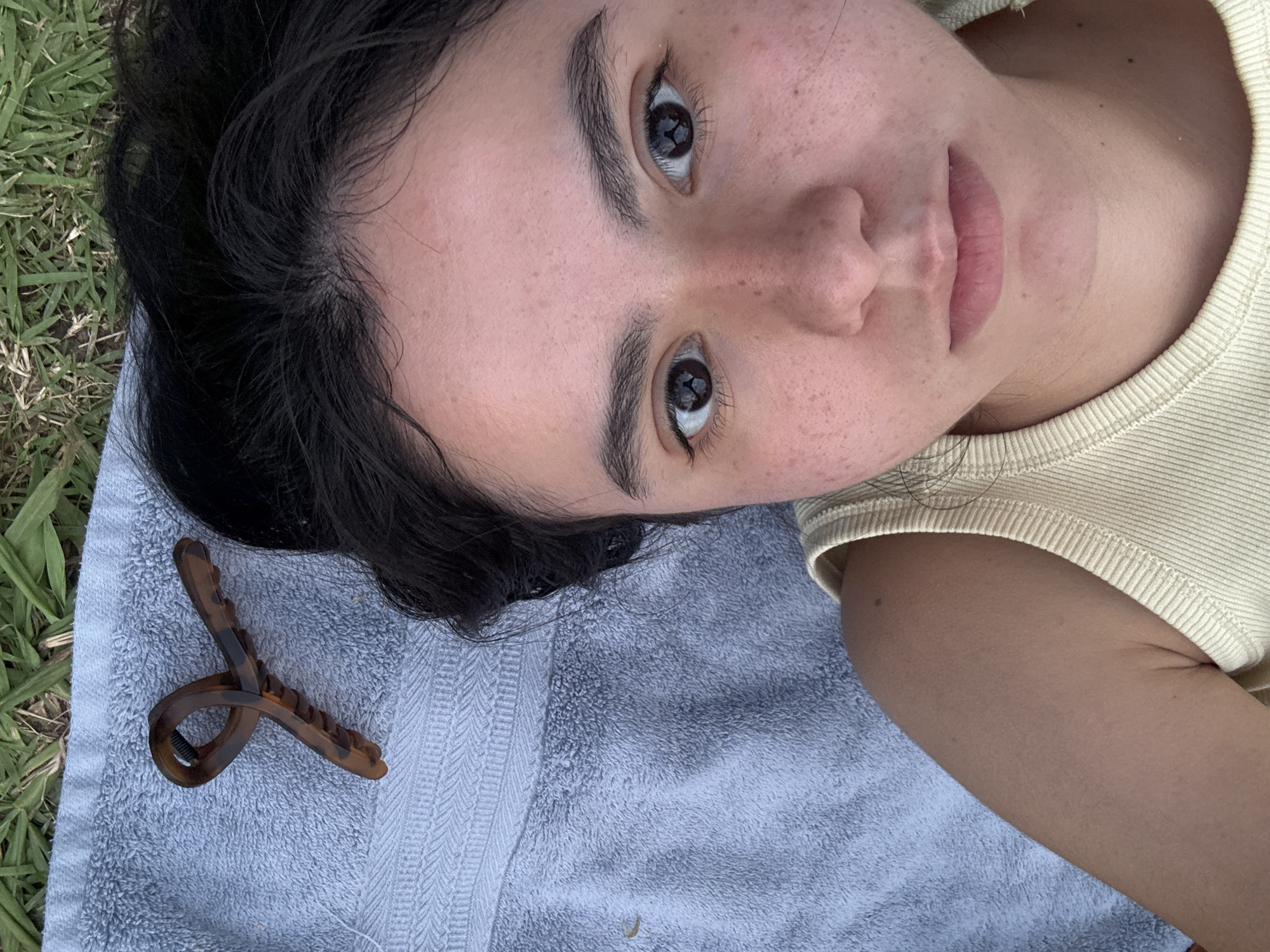 The image shows a person lying on a light blue towel. The person has dark hair and is wearing a light yellow top. A brown hair clip is lying on the towel near their head. The background includes grass. The image does not contain enough information to determine a specific location within the city of Austin, United States of America.