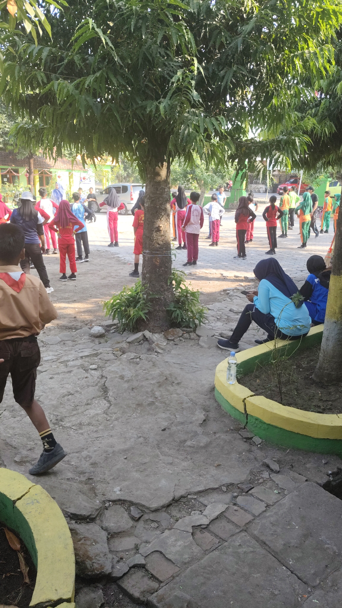 In Banjarsari, Indonesia, a group of children in school uniforms are participating in an outdoor activity, possibly physical education class, under the shade of a large tree. The children are wearing a variety of colored uniforms, including red, white, and green. Some children are standing in groups, while others are seated. One child in the foreground is wearing a tan uniform with a red and white scarf, suggesting a scouting or cadet uniform. The ground is paved with concrete and some areas show signs of wear and tear, with cracked pavement and exposed soil. The environment appears to be a schoolyard or park, with buildings and parked vehicles visible in the background. The lighting suggests it is daytime, with sunlight filtering through the leaves of the tree.