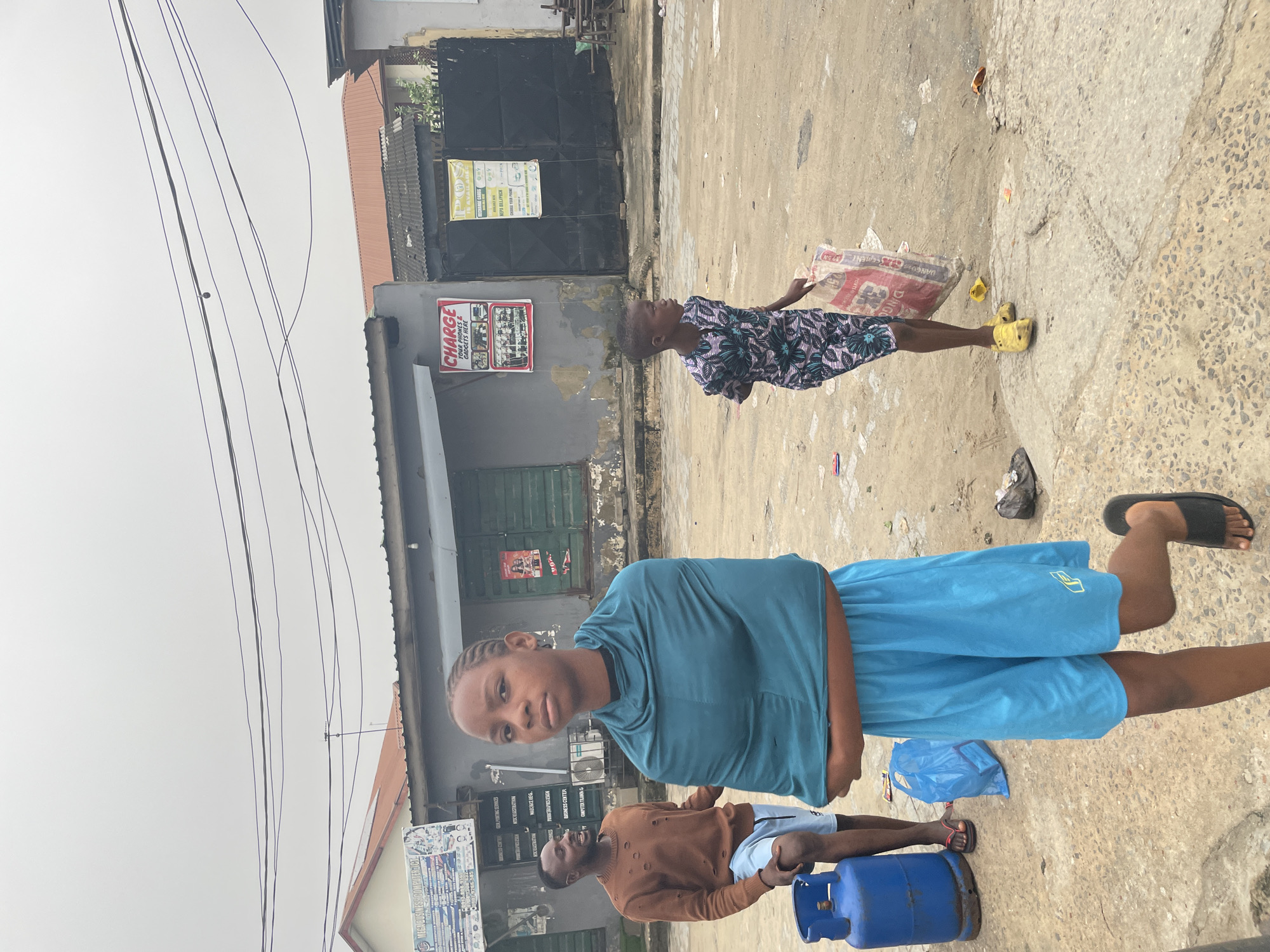 This is an outdoor street scene in Lagos, Nigeria, featuring three individuals and several commercial buildings under an overcast sky.
In the foreground, a young Black female, approximately early teens, stands center-left, looking directly at the camera with a neutral expression. She has braided hair and wears a teal-colored fabric draped over her shoulders and torso, light blue athletic shorts with a yellow logo, and black flip-flops. Her right foot is slightly raised.
To her left, a young adult Black male with a beard stands with his mouth open, seemingly laughing or speaking, looking towards the left. He wears a light brown, distressed long-sleeved shirt and light blue shorts with white trim, along with dark flip-flops. Beside him on the ground is a blue gas cylinder. A crumpled blue plastic bag lies on the ground between him and the central girl.
To the right, a young Black child, appearing pre-teen, walks leftward across the frame, carrying a large white and red sack (partially identifiable as "DANGOTE CEMENT") in their right hand. The child wears a patterned outfit with blue, pink, and black designs and yellow slip-on shoes.
The ground is an uneven mix of dirt, gravel, and cracked concrete with scattered litter. In the background, several single-story commercial buildings line the street. Signs on these buildings advertise services such as "MEGA BUSINESS COMPUTER ICT" (offering computer training, repairs, graphic design, etc.), "CHARGE YOUR PHONES & GADGETS HERE," and "POS IS AVAILABLE HERE" (for recharge cards, bill payments, phone charging). Some buildings have dark green shutters, security gates, and an outdoor air conditioner unit is visible. Numerous black electrical wires crisscross the frame above the buildings.
