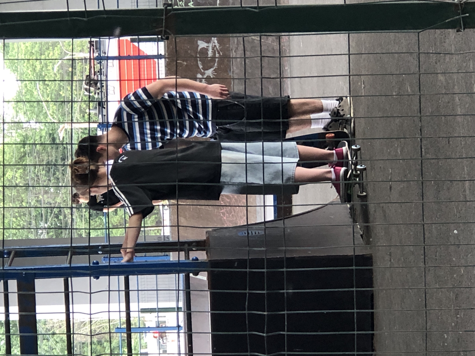 In Saint Petersburg, Russia, behind a chain-link fence, two young men are visible in a skate park. The man on the left wears a black t-shirt with white stripes on the sleeves and light-colored shorts. He is standing with one foot on a skateboard, his left hand extended forward. The man on the right wears a blue and white striped t-shirt and black shorts, with white socks and dark sneakers. He is standing next to the first man, facing forward. Both men appear to be teenagers or young adults. The background reveals trees, a red and white structure possibly a building or train car, and a skate park ramp structure. The ground is paved and appears to be asphalt. The overall lighting suggests it is daytime.