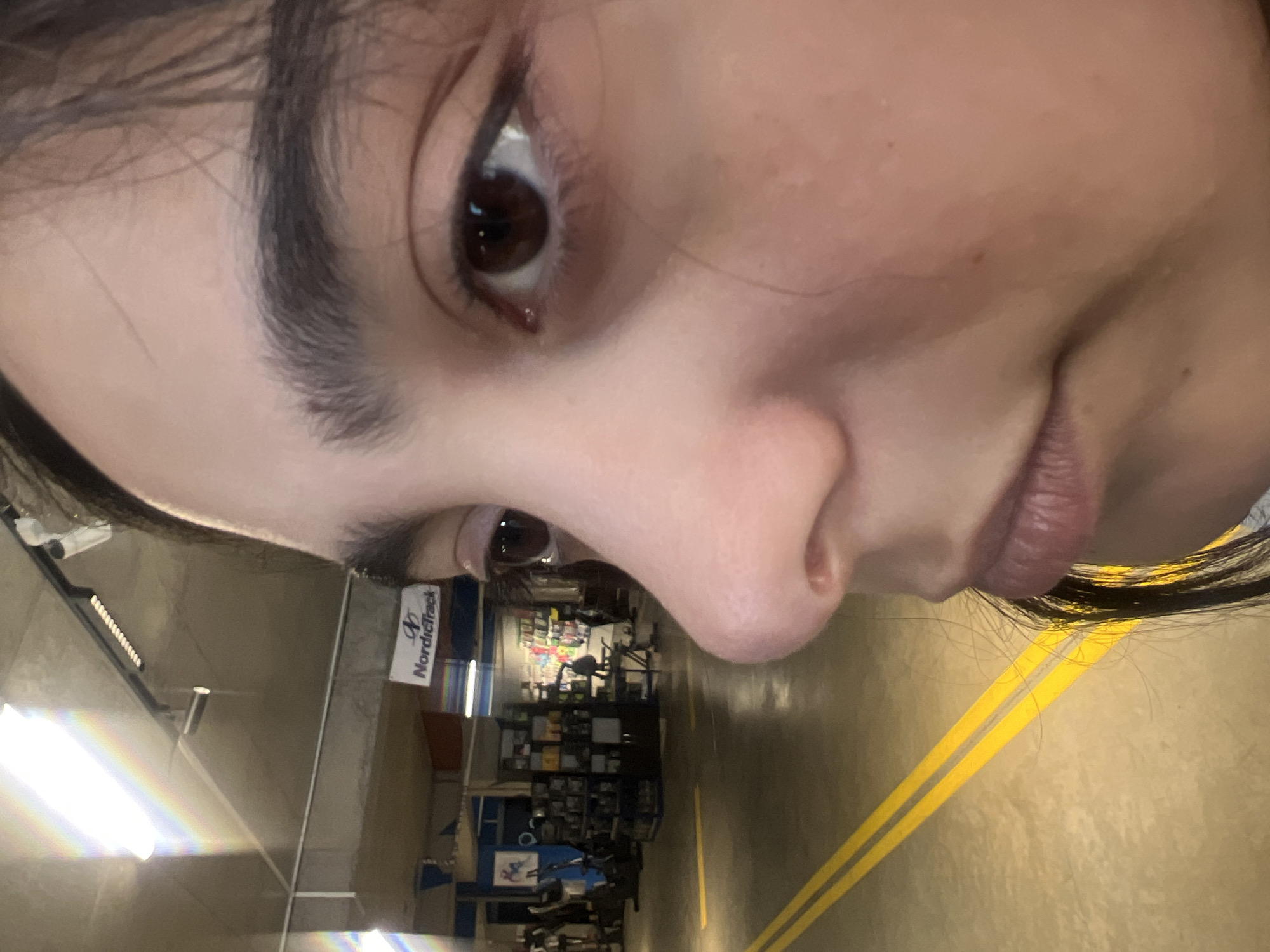 A selfie of a person with brown eyes, taken from a close and angled perspective. The person has dark hair and light skin. The background appears to be inside a store, possibly a sporting goods store, with rows of exercise equipment visible. A "NordicTrack" sign is visible, along with yellow floor markings. There is no information in the image to indicate that it was taken in the city of Tbilisi, Georgia.