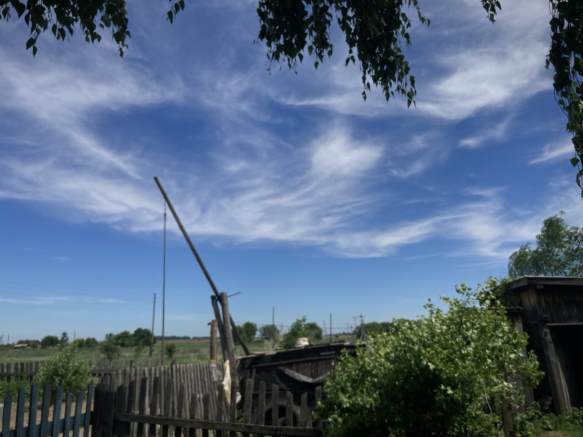 The image shows a view of a rural area under a partly cloudy blue sky, with foliage from trees in the foreground obscuring the top edges of the frame. A weathered wooden fence runs across the lower portion of the image, with some sections appearing dilapidated. Behind the fence, there is a rudimentary wooden structure, possibly an outbuilding or shed, with a dark opening. Further in the background, a flat, grassy landscape stretches to the horizon, dotted with scattered trees and some buildings that suggest a small village or settlement. Several thin poles, some appearing to be utility poles or supports, are visible against the sky. The scene evokes a sense of a rustic, perhaps underdeveloped, environment. There are no people visible in the image. The location is specified as Rakity, Russia.