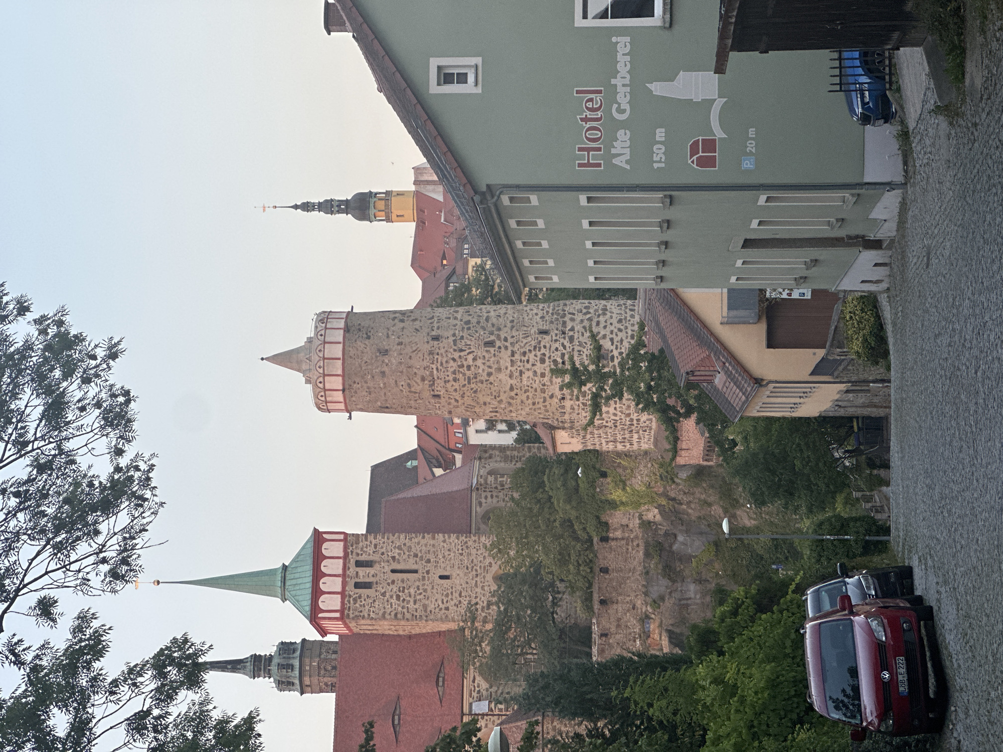 This image depicts a view of the historic city of Bautzen, Germany, looking uphill on a cobblestone street lined with buildings. In the background, several prominent medieval towers and structures of Bautzen's altstadt (old town) are visible, including the Reichenturm and the Ortenburg castle complex with its distinctive red roofs and stone towers, some adorned with white decorative elements and red accents. The sky is overcast and pale. In the foreground, a light green building on the right side of the street displays signage for "Hotel Alte % Gerberei" with an indication of parking at "150 m" and "P 20 m". A dark blue car is parked in front of this building. On the left side of the street, a red car and a darker car are parked on the uphill slope. Lush green trees and vegetation are interspersed throughout the scene, adding a natural element to the urban landscape. The overall impression is one of an old European city with well-preserved historical architecture.