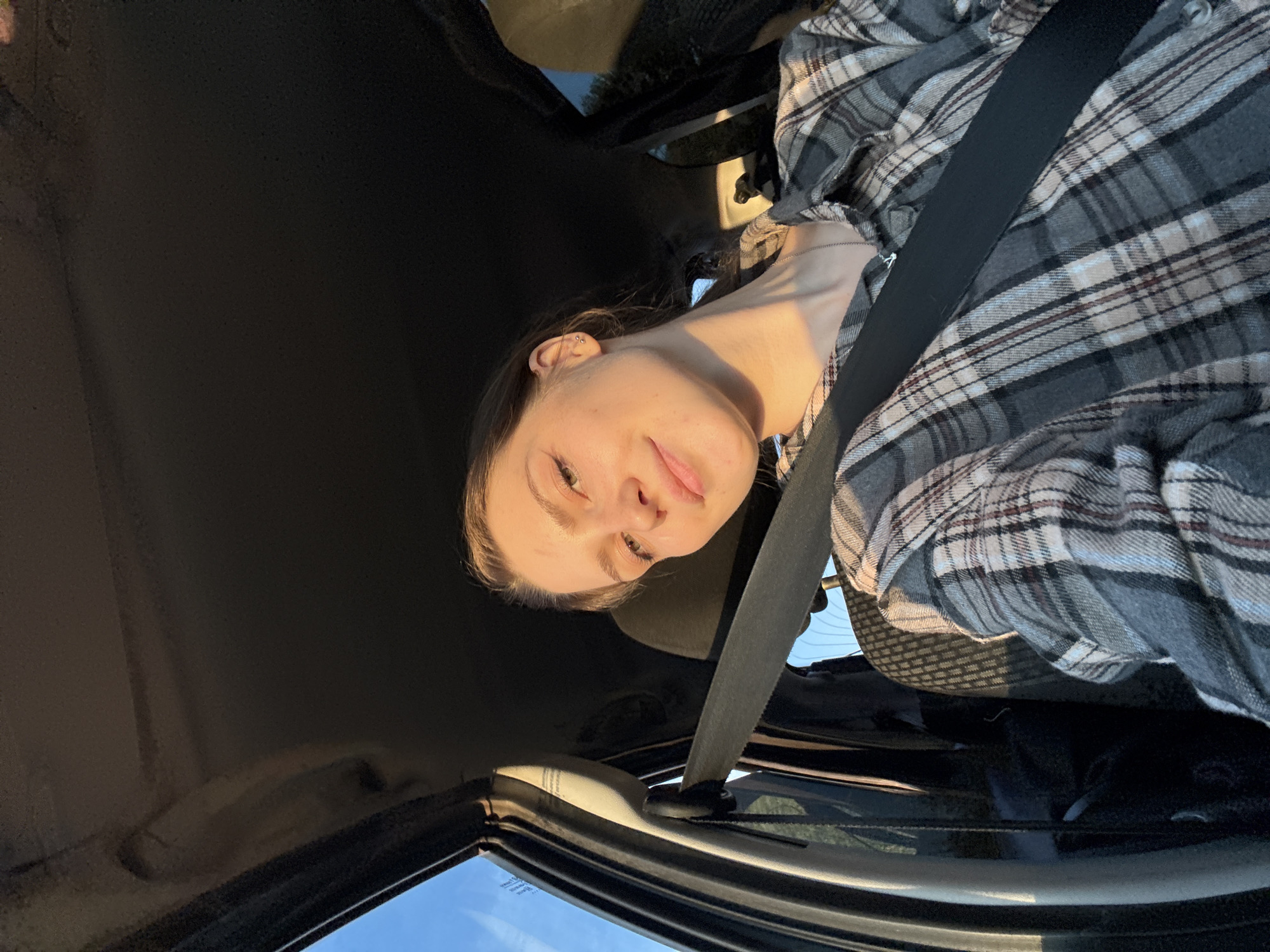 A young woman with brown hair pulled back is sitting in the passenger seat of a car, wearing a plaid shirt and a seatbelt. She is looking towards the camera with a slight smile, illuminated by the golden light of the setting sun, which casts a warm glow on her face and neck. The interior of the car is visible, with the dark headliner and dashboard in the background, and the blue sky and faint treeline visible through the window on the left. The image was taken in Дресвяново, Russia, likely during a car ride at sunset.